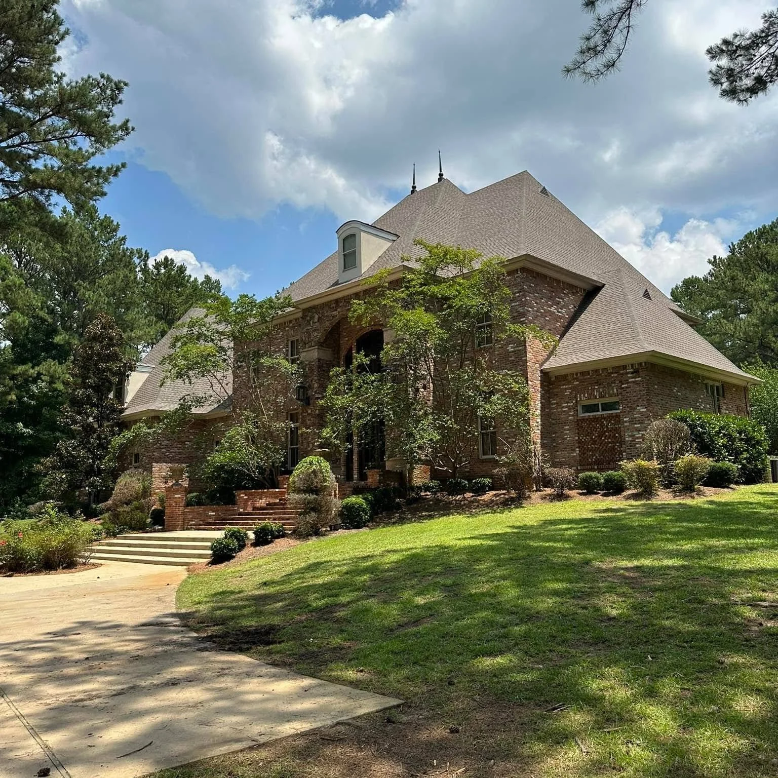 Large brick house in a lush green yard with trees, steps, and a dirt driveway, under a partly cloudy sky.