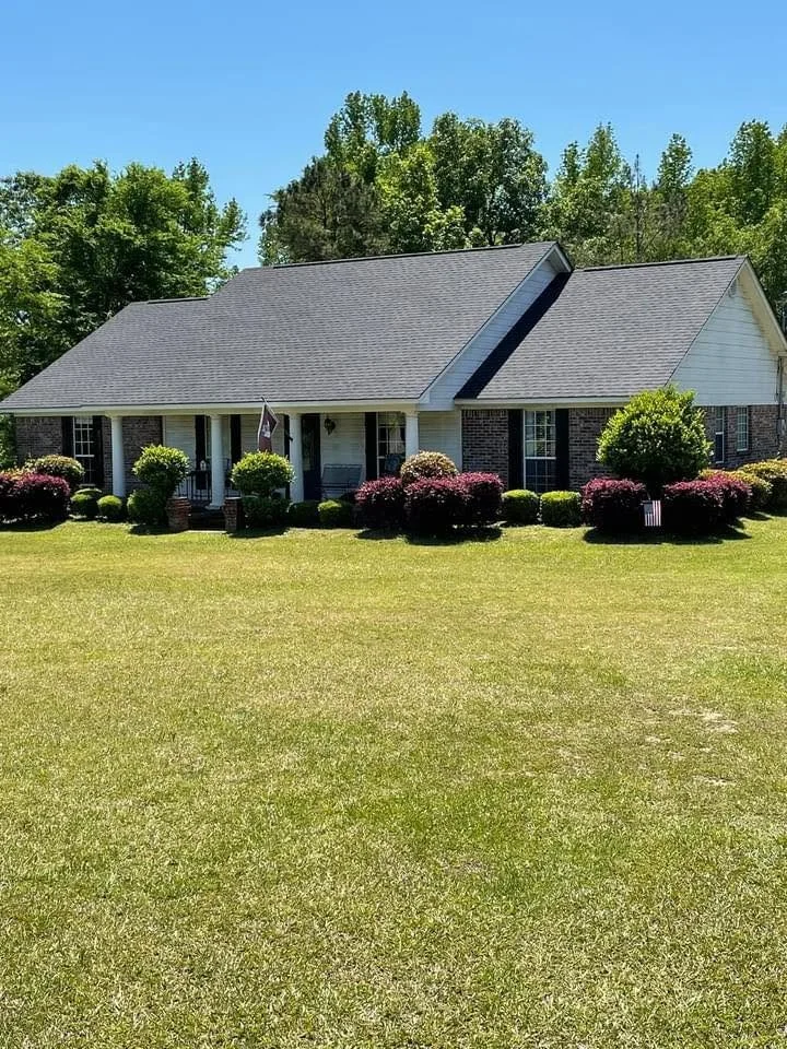 A single-story house with a gray shingled roof and white exterior walls, surrounded by neatly trimmed bushes and a well-maintained grassy lawn, with trees and a clear blue sky in the background.