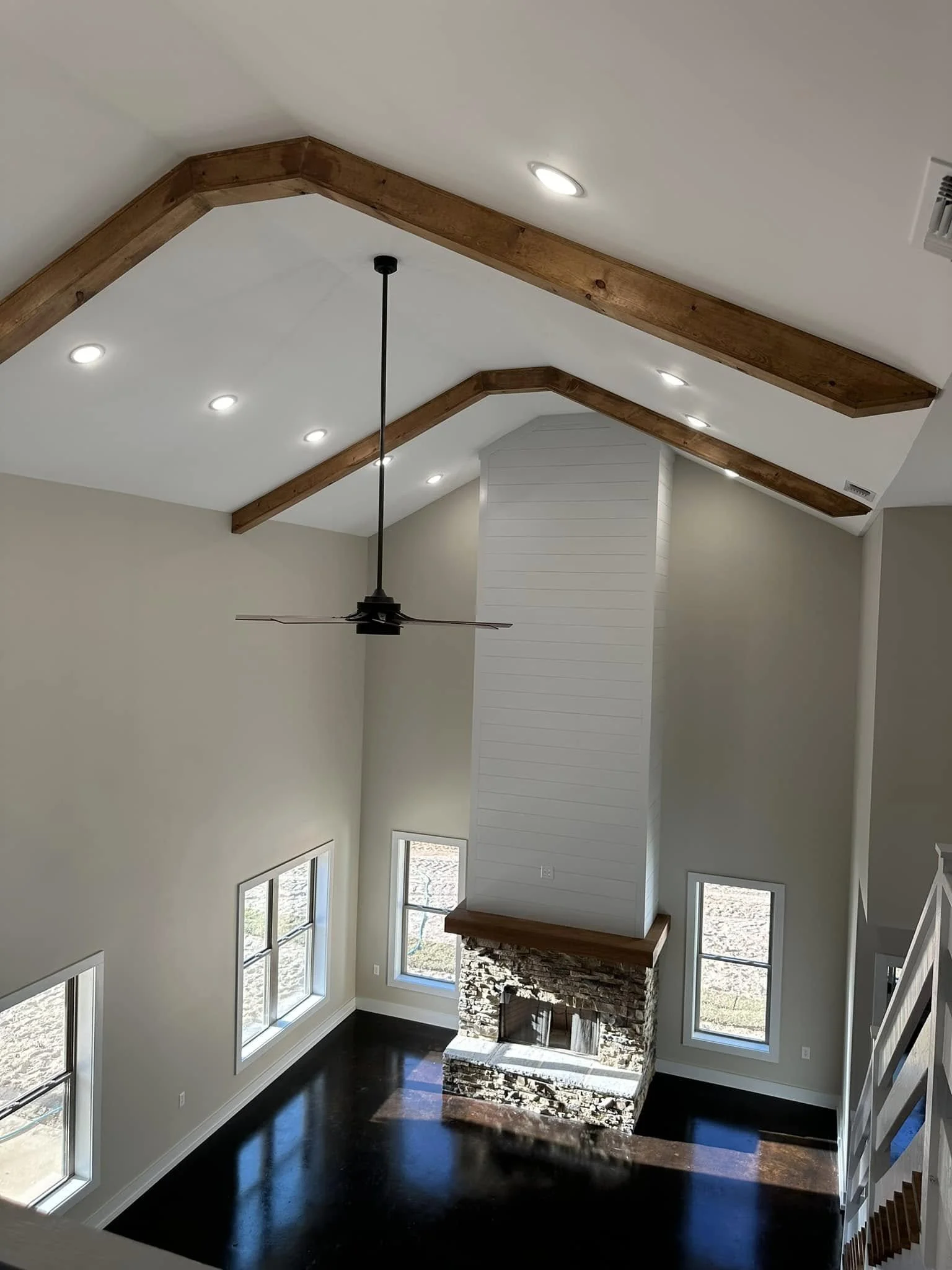 A spacious living room with high vaulted ceiling that has elegant wooden beams, a stone fireplace, multiple windows allowing natural light, and a ceiling fan, with dark hardwood floors and light-colored walls.