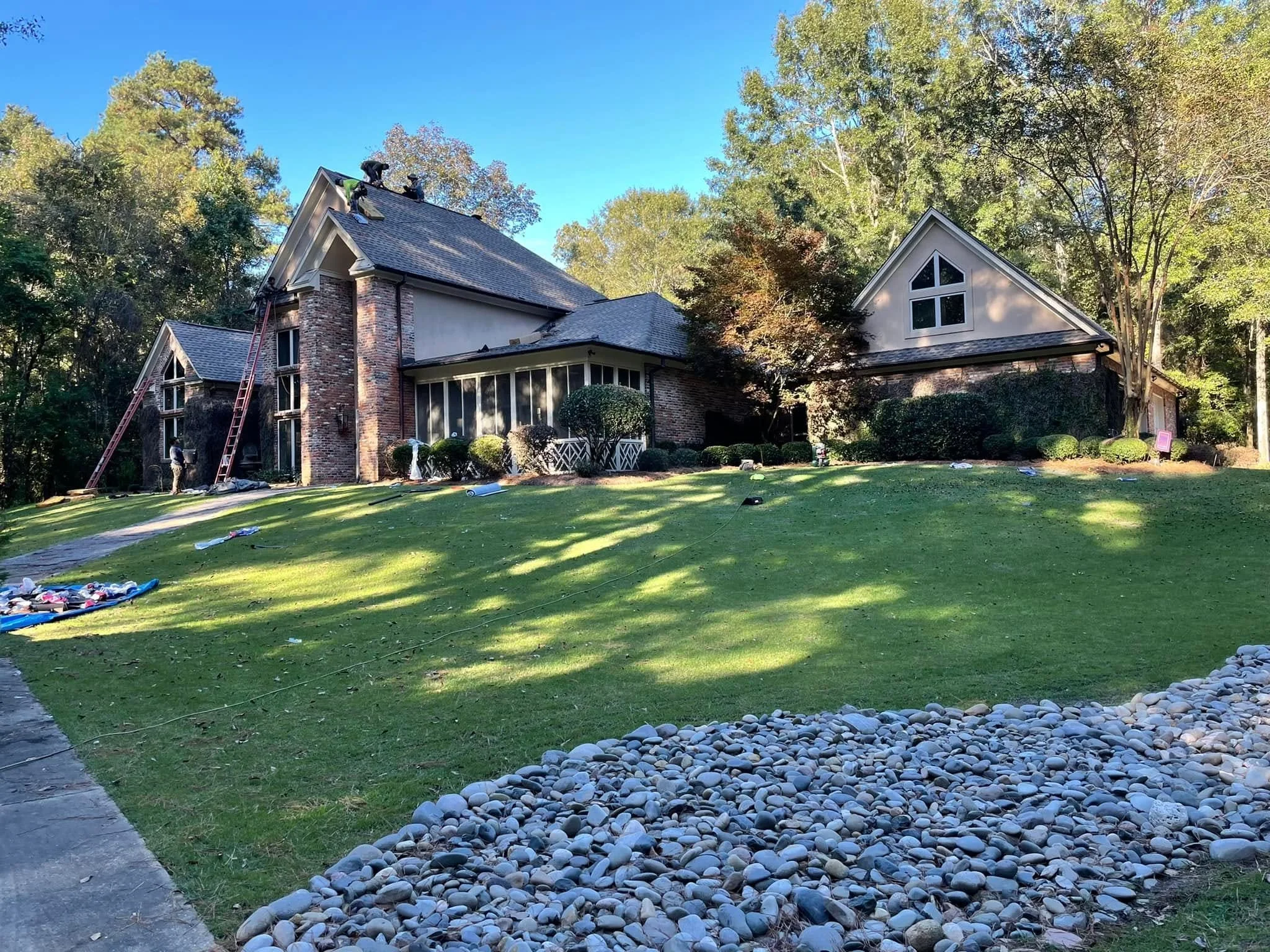 Workers on the roof of a two-story brick house replacing shingles during the daytime with a background of green trees and a clear blue sky.
