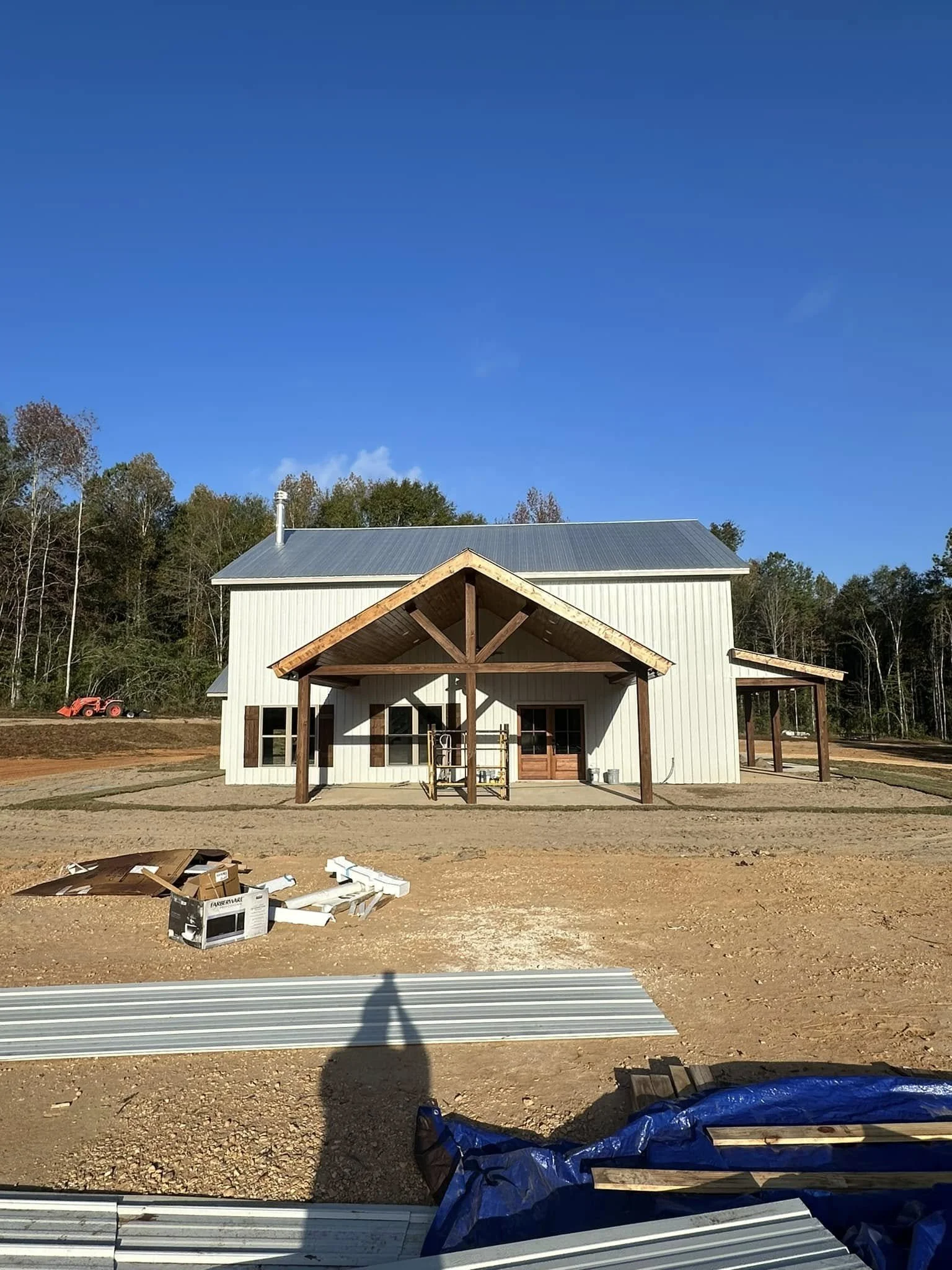 A building under construction with a metal roof, a porch with wooden beams, and surrounded by trees and construction materials on a clear day.