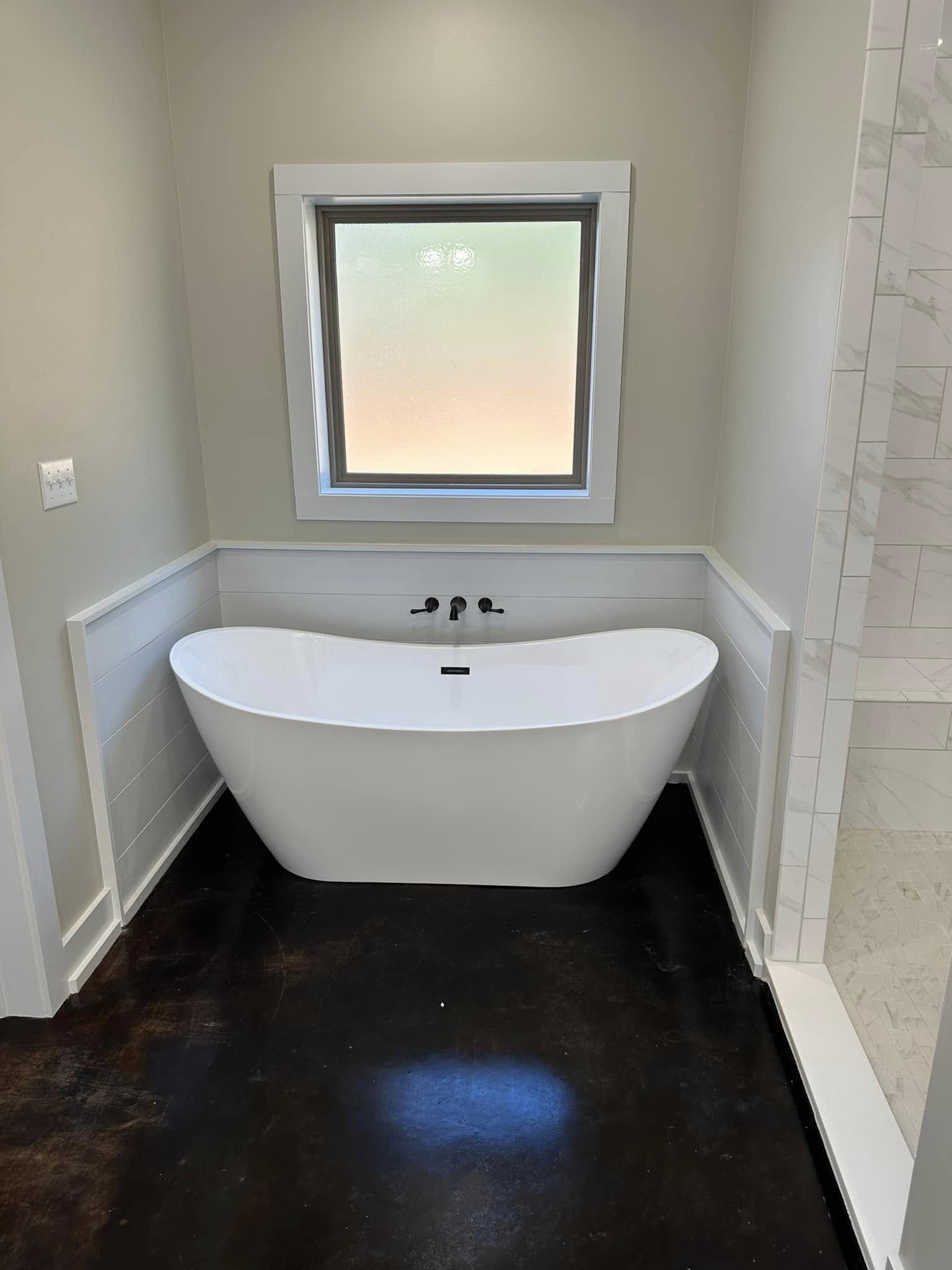 A modern bathroom with a freestanding white bathtub beneath a frosted window, surrounded by beige walls, white trim, and dark wooden flooring.