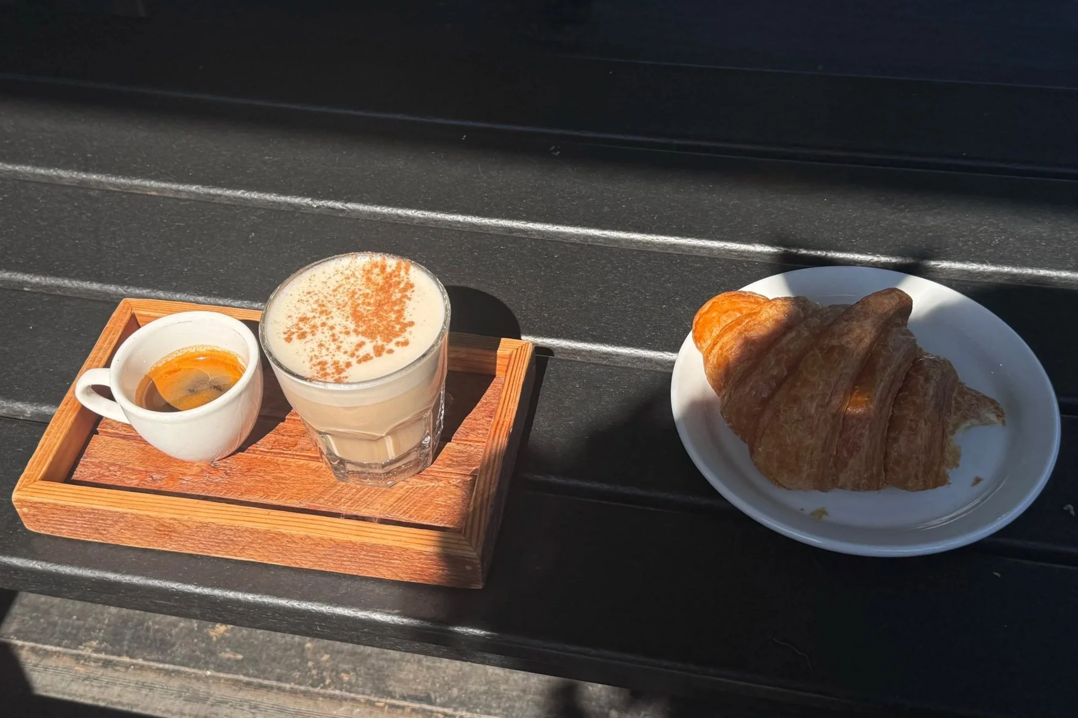 A wooden tray holding a small cup of coffee, a glass of latte with a foam topping, and a white plate with a croissant. The tray is placed on a black surface outdoors.