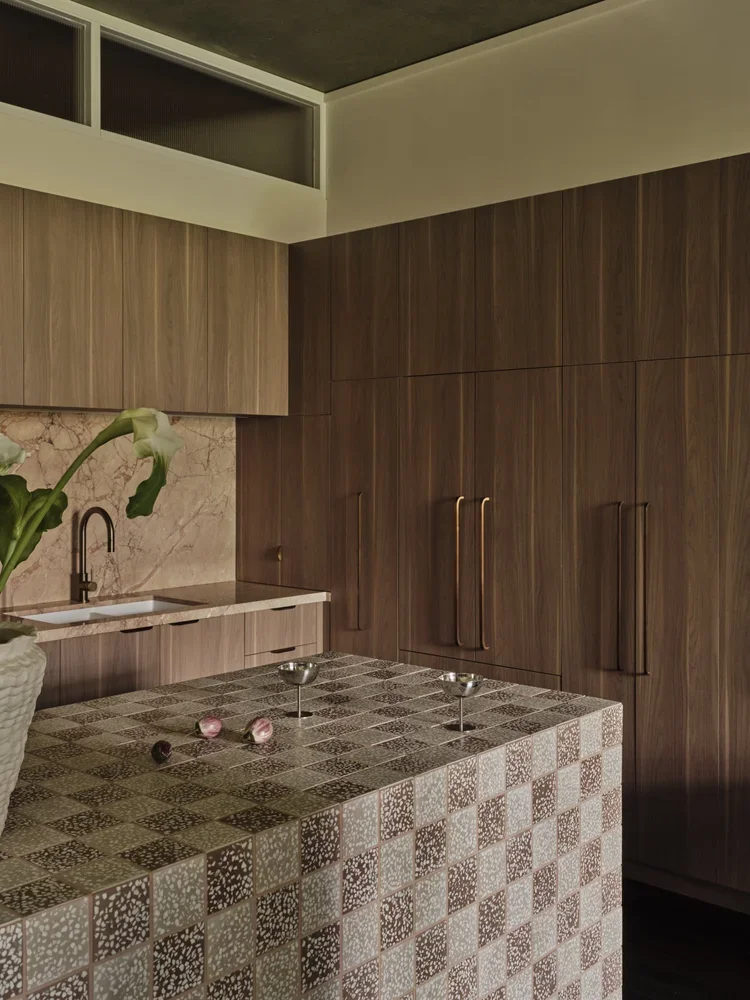 Modern kitchen with wooden cabinets, a tiled island, and a marble backsplash. A plant with green leaves and white flowers is on the counter.