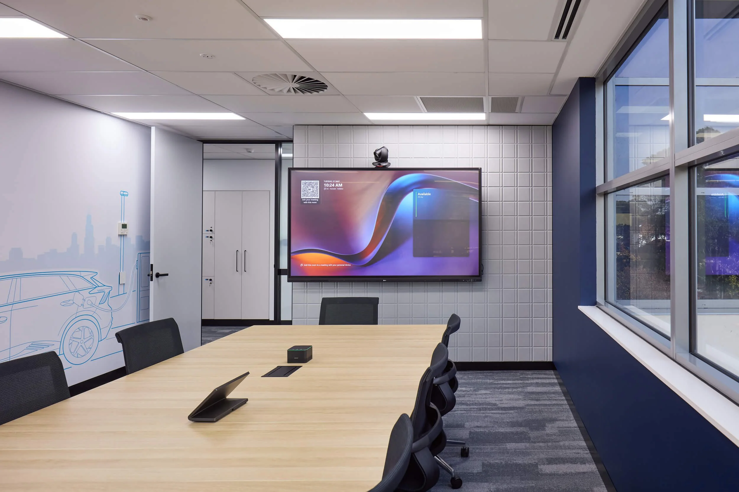 A modern conference room with a large wooden table, black chairs, a digital display on the wall, and large windows showing outside scenery.