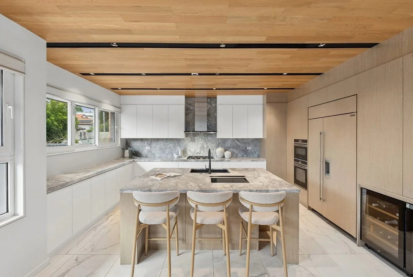 Modern kitchen with light wood cabinets, marble countertops, and a wooden ceiling, featuring a central island with seating, stainless steel appliances, and a large window.