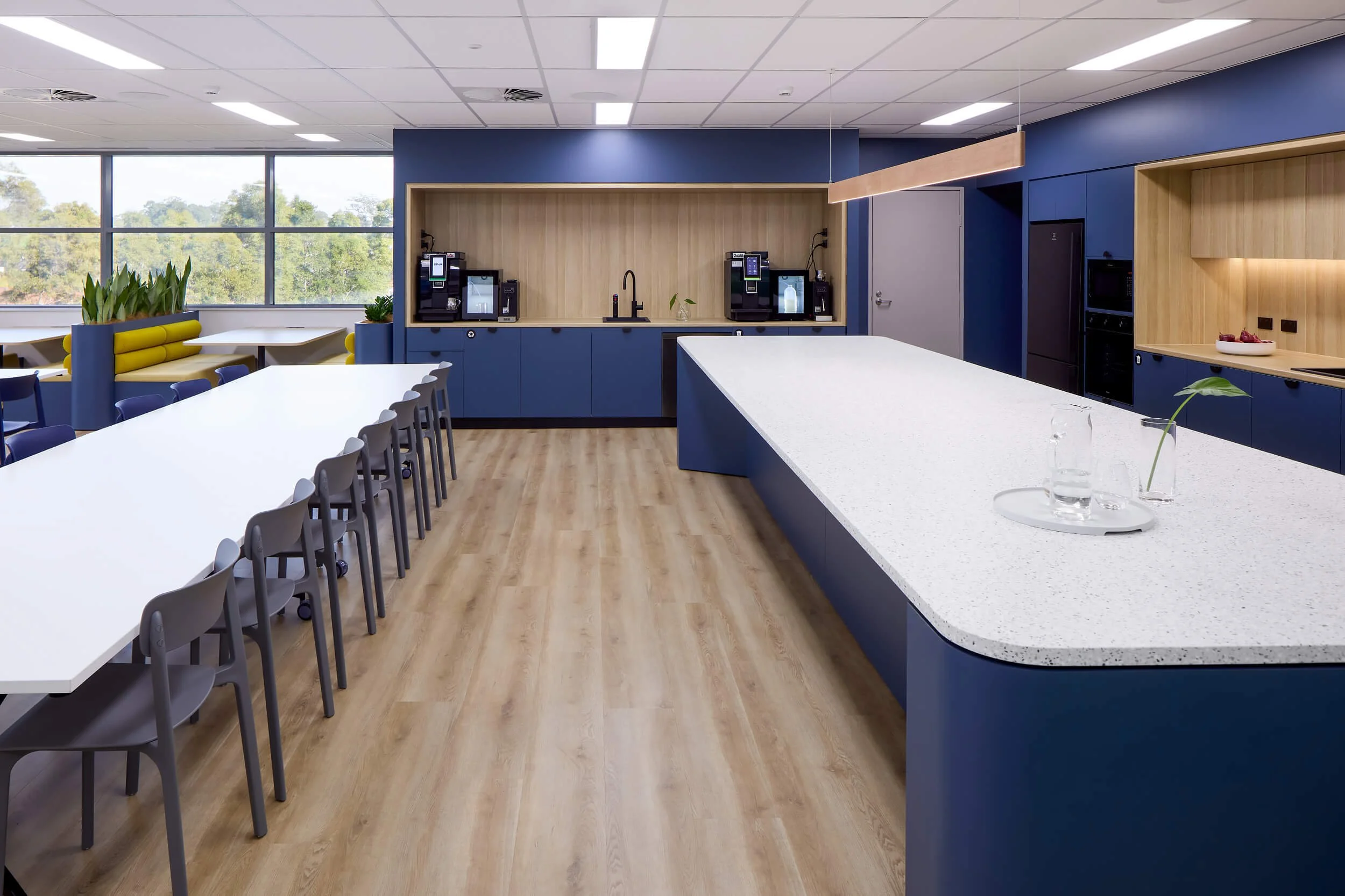 Modern break room with large windows, blue walls, white countertop, coffee machines, plants, and seating area with yellow cushions.