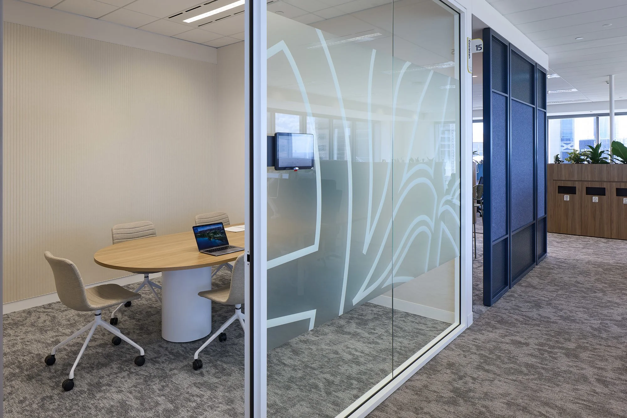 Modern office conference room with a round table, four beige chairs with wheels, a laptop, a glass meeting room with frosted designs, and blue acoustic panels. There are large windows with cityscape views, a wooden planter with green plants, and a gray carpeted floor.