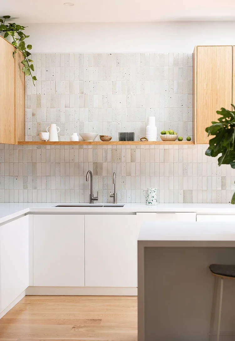 Modern kitchen with white cabinets, wooden open shelf with white and neutral-colored dishware, backsplash with small square tiles, stainless steel faucet, potted plants, and a few green apples in a bowl.