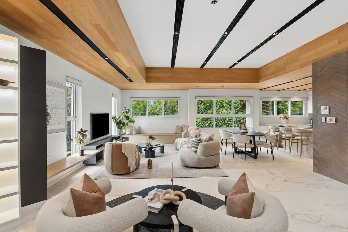 Spacious modern living room with large windows, beige sofas, round chairs, a flat-screen TV, and a mix of wood and marble flooring and ceilings.