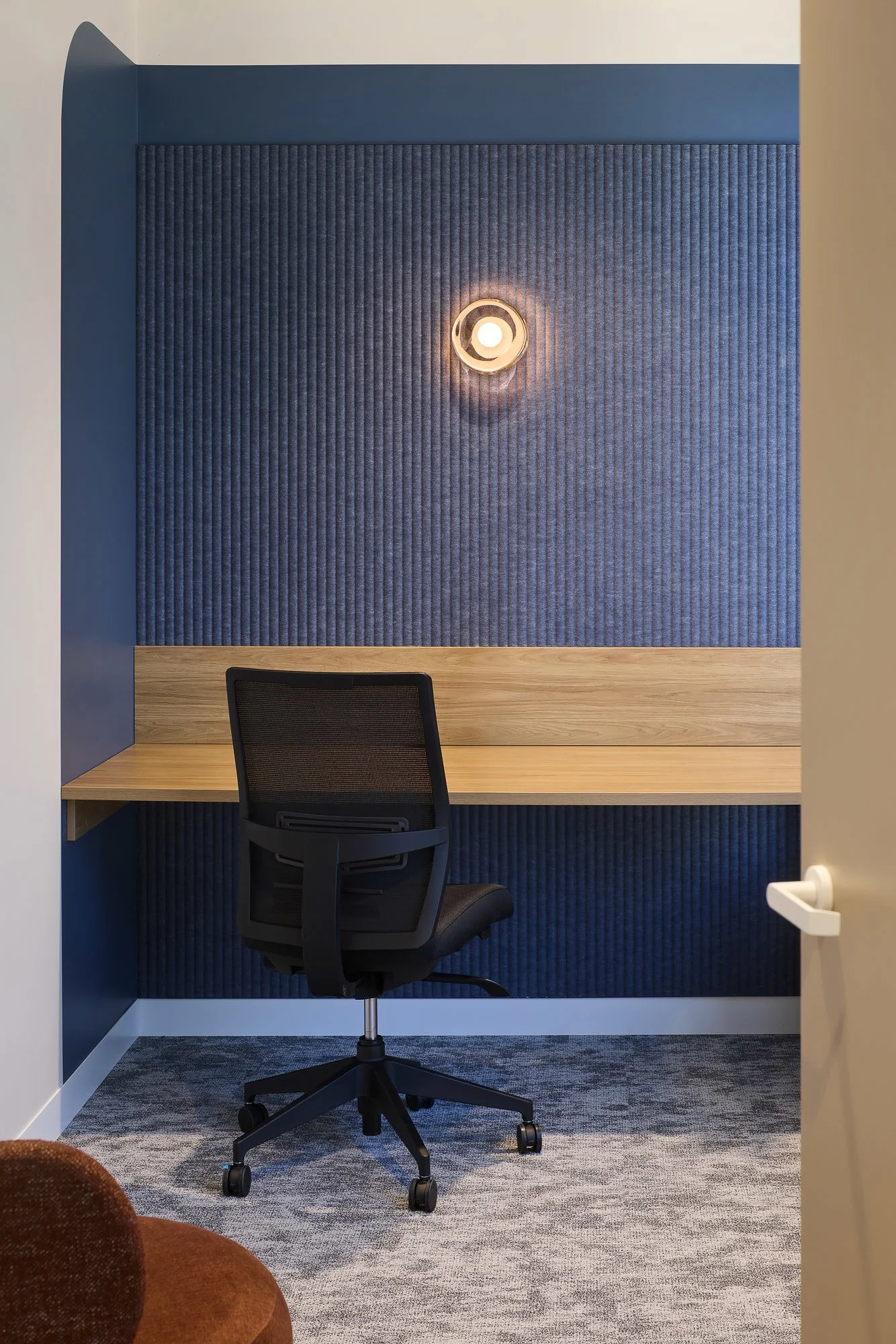 An office workspace with a black ergonomic chair, a wooden desk, a blue acoustic panel on the wall, and a round wall light fixture in the center of the panel.