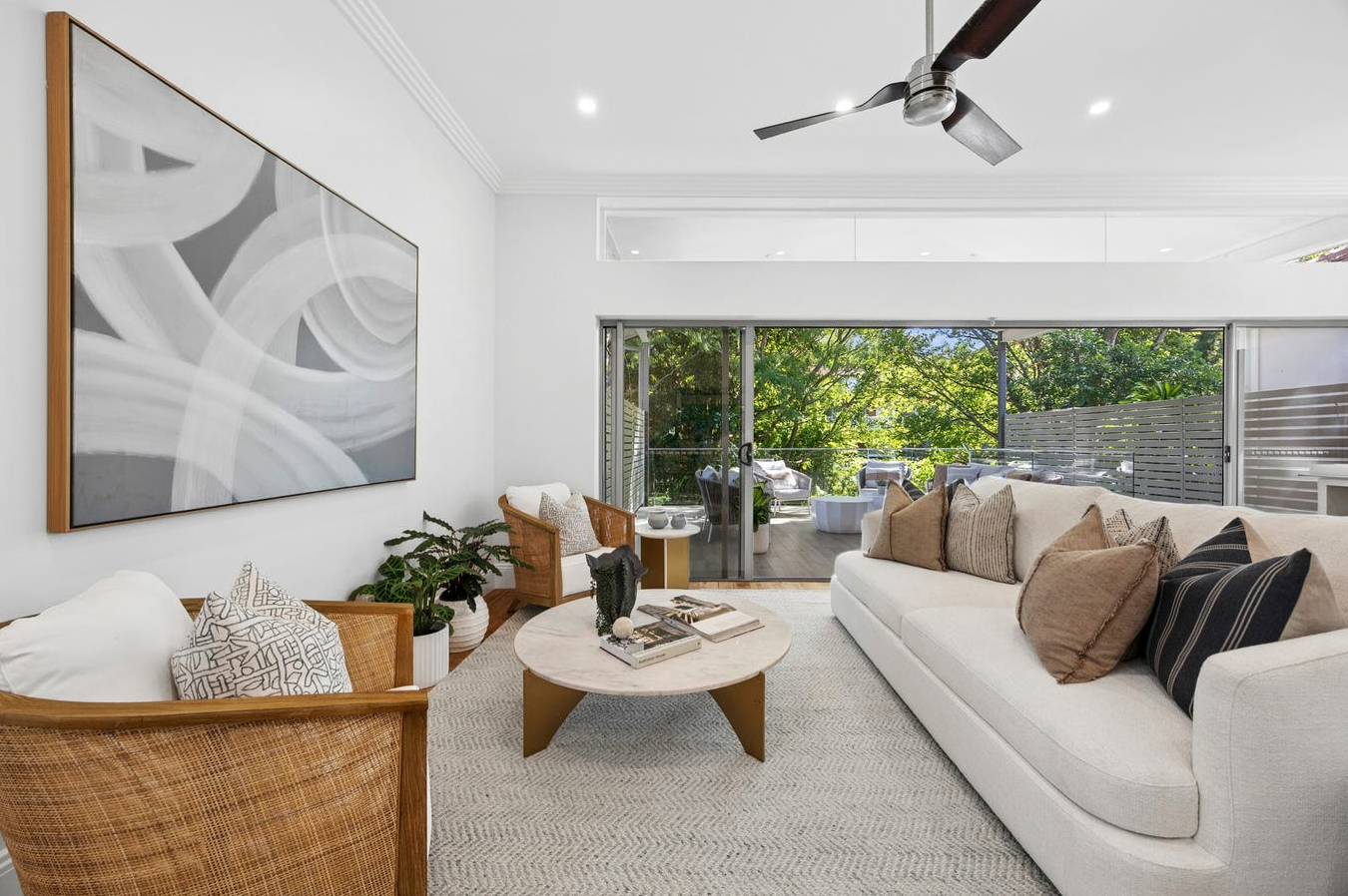 Living room with white walls, large abstract black and white art, wicker chairs, white sofa with beige and black pillows, coffee table with books, potted plants, and a sliding glass door leading to a patio with outdoor seating and green trees.