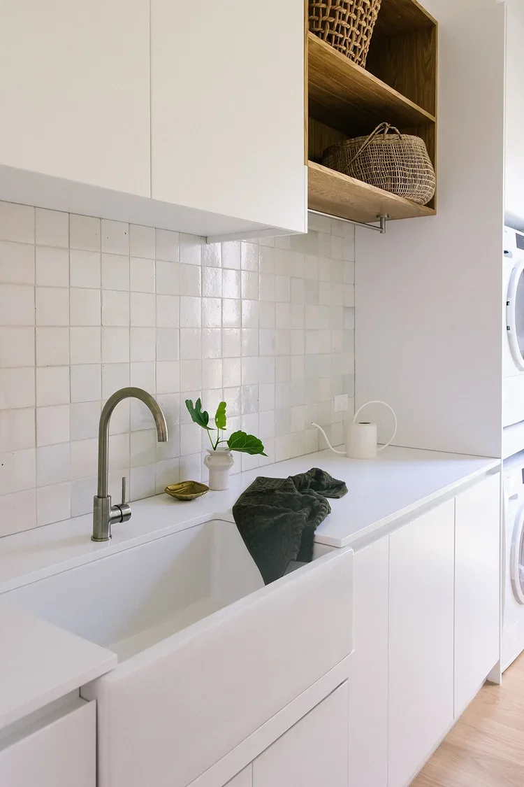 Modern laundry room with white cabinets, a white sink, a black towel, potted green plant, soap dish, watering can, open wooden shelves with baskets, and a washing machine in the background.