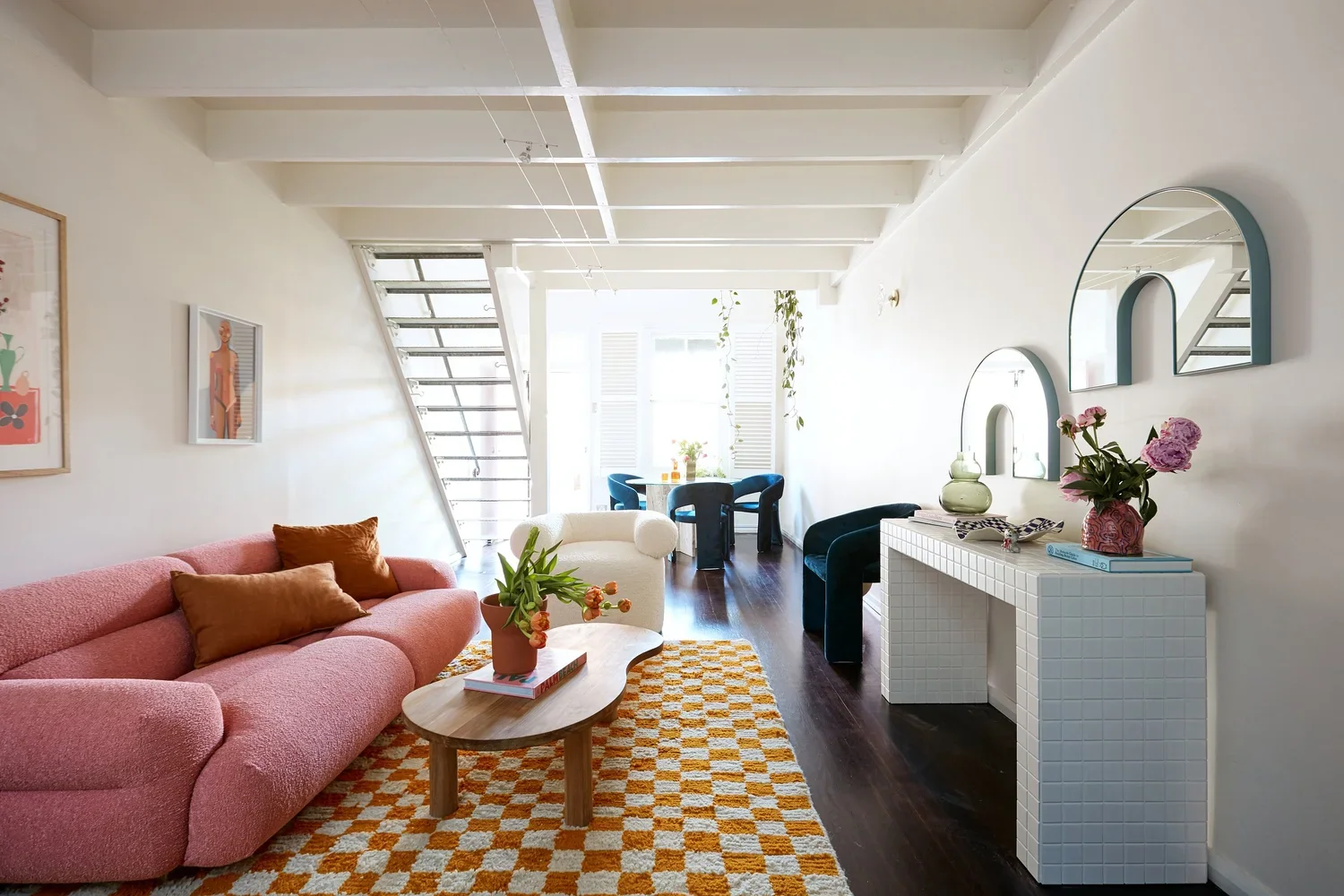 Bright living room with pink sofa, wooden coffee table, and various decorative items. Dining area with blue chairs and large windows at the back, natural light, plants, and white walls with round mirrors.