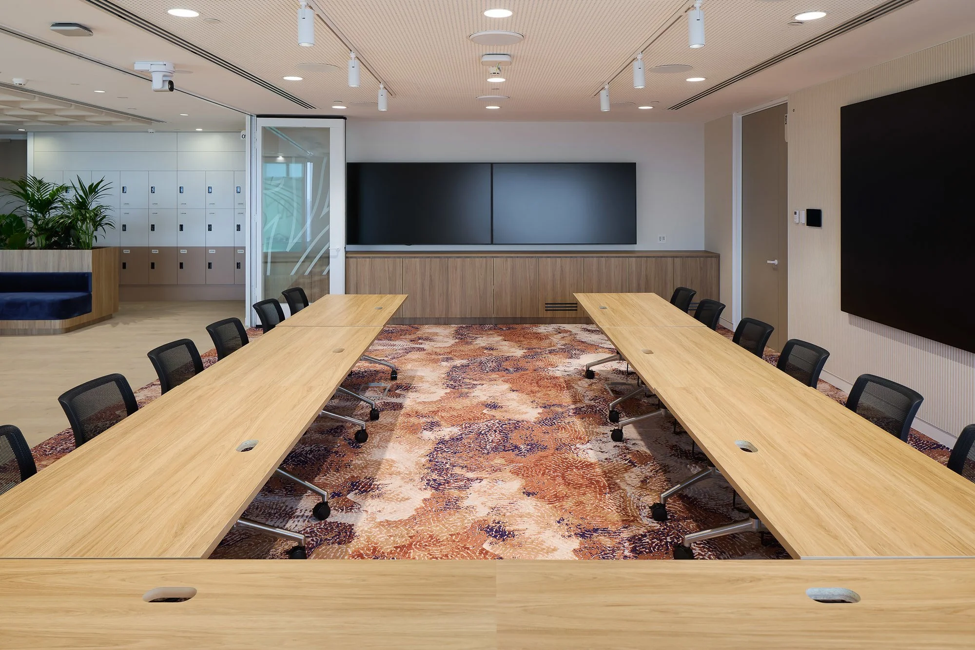 Modern conference room with a large wooden table, black mesh chairs, a hanging blackboard, a colorful area rug, lockers, plants, a glass door, and a large black wall panel.