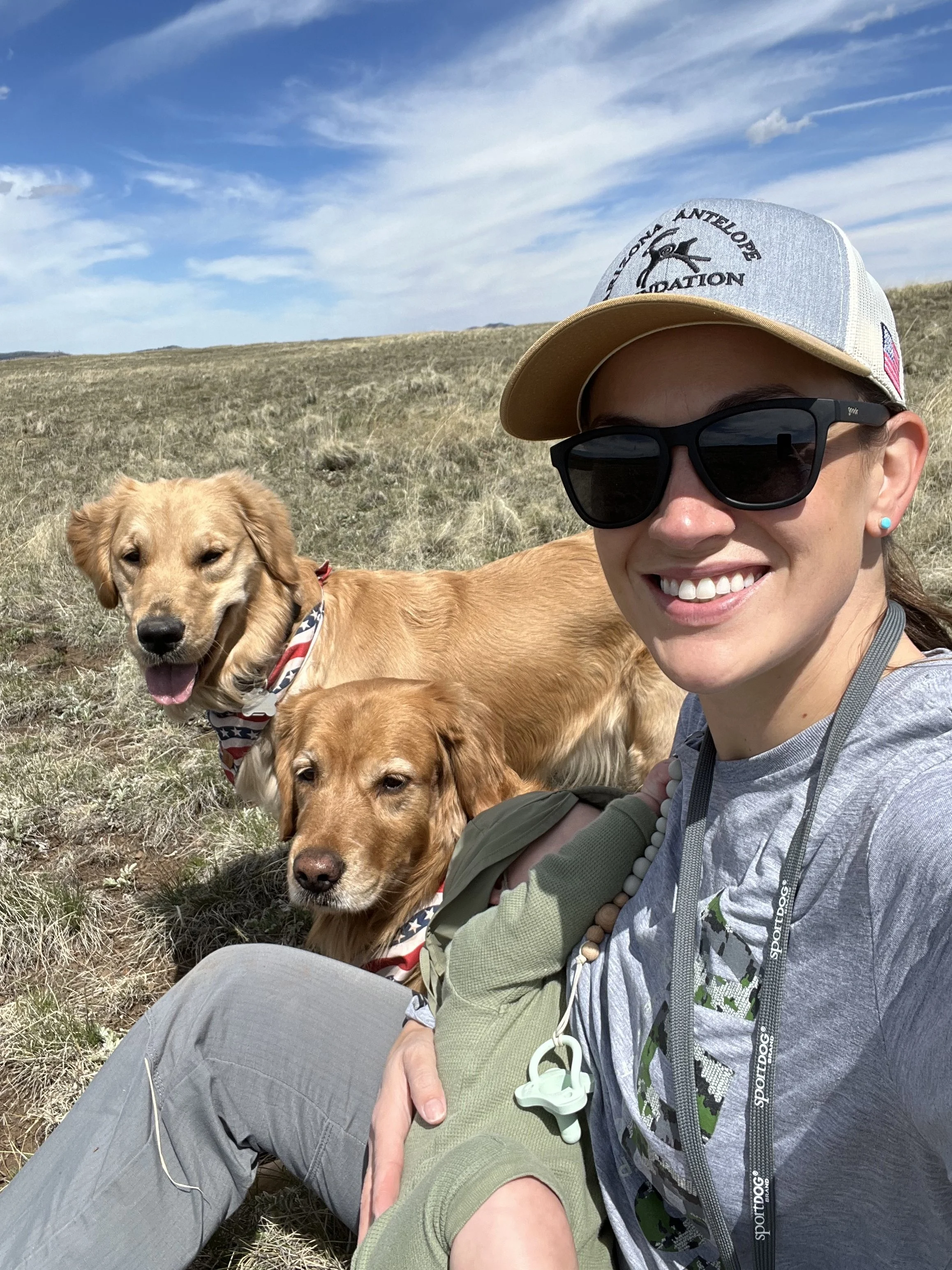 A woman wearing sunglasses and a cap with a logo, sitting outdoors in a grassy field on a sunny day, with three friendly golden retriever dogs, one of which is wearing a patriotic bandana.