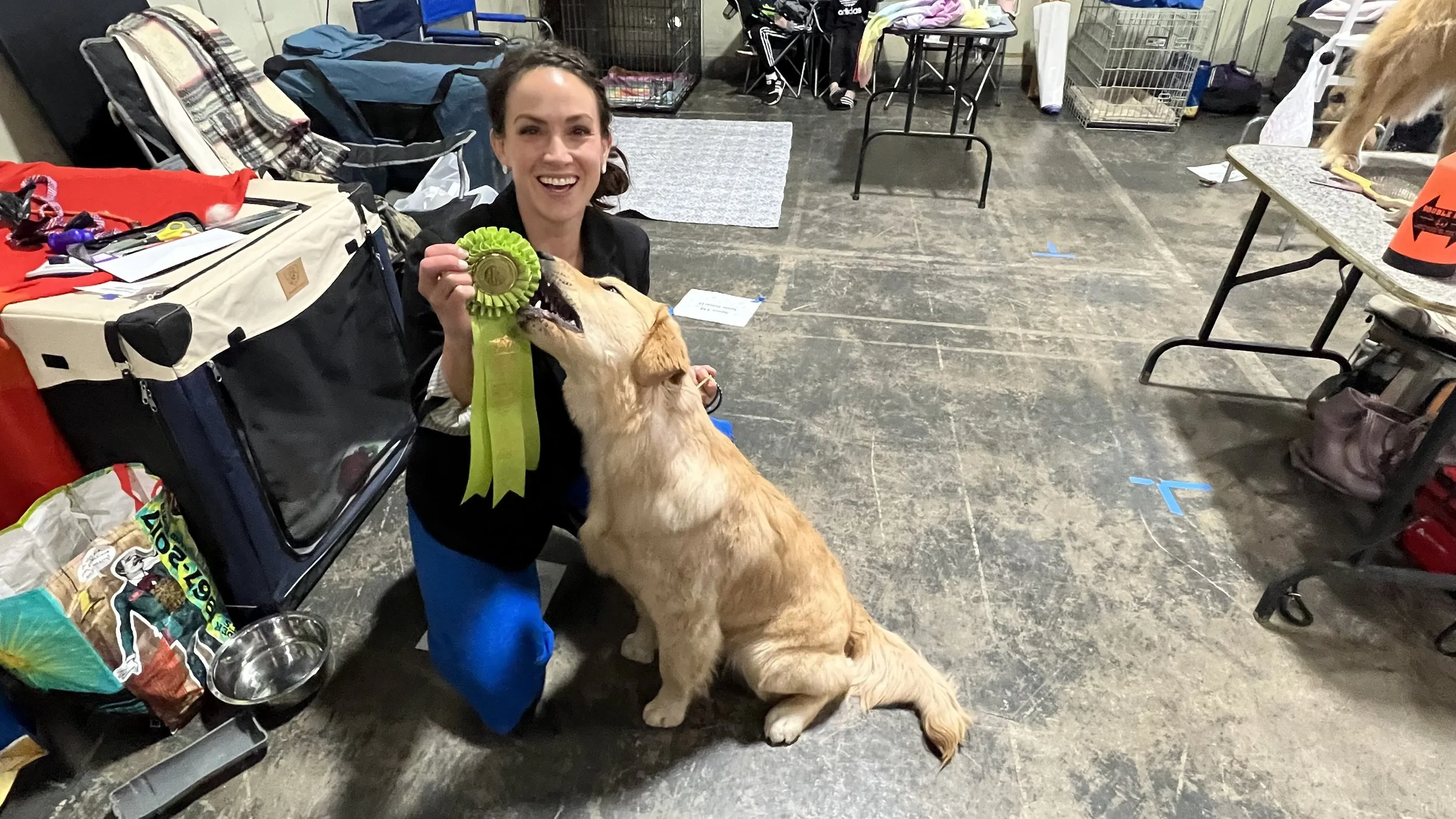 A woman smiling and holding a yellow ribbon award with her hand, next to a golden retriever dog at an indoor event with tables, chairs, and dog supplies.