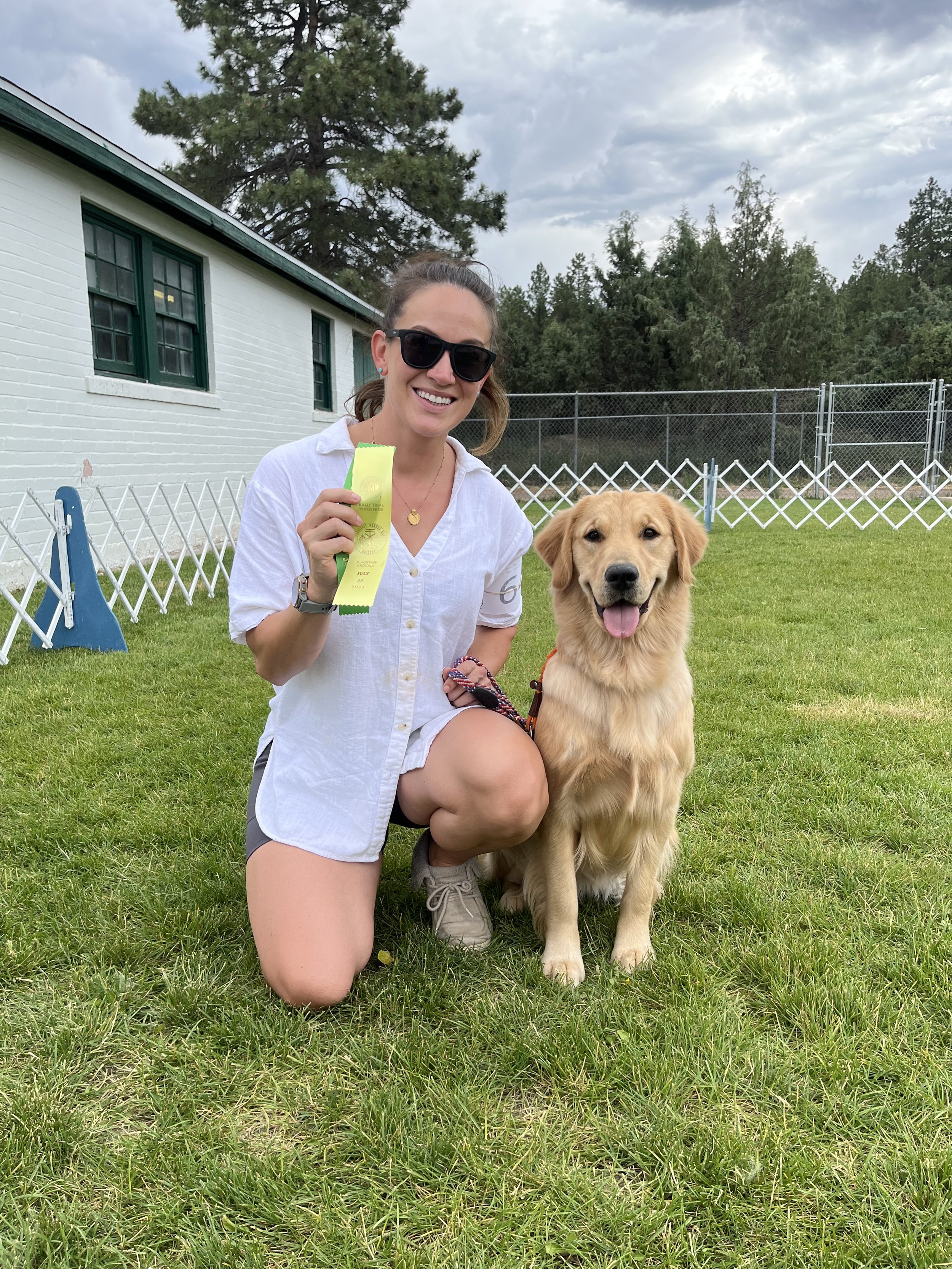 A woman wearing sunglasses and a white shirt kneels on the grass next to a golden retriever dog. She is holding a yellow and green ribbon and smiling. The dog is sitting with its tongue out. There is a white building with green window frames and a ch