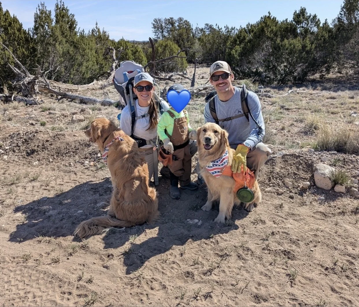 Two adults with a young child and three dogs outdoors in a desert-like area with bushes and rocks, smiling at the camera.