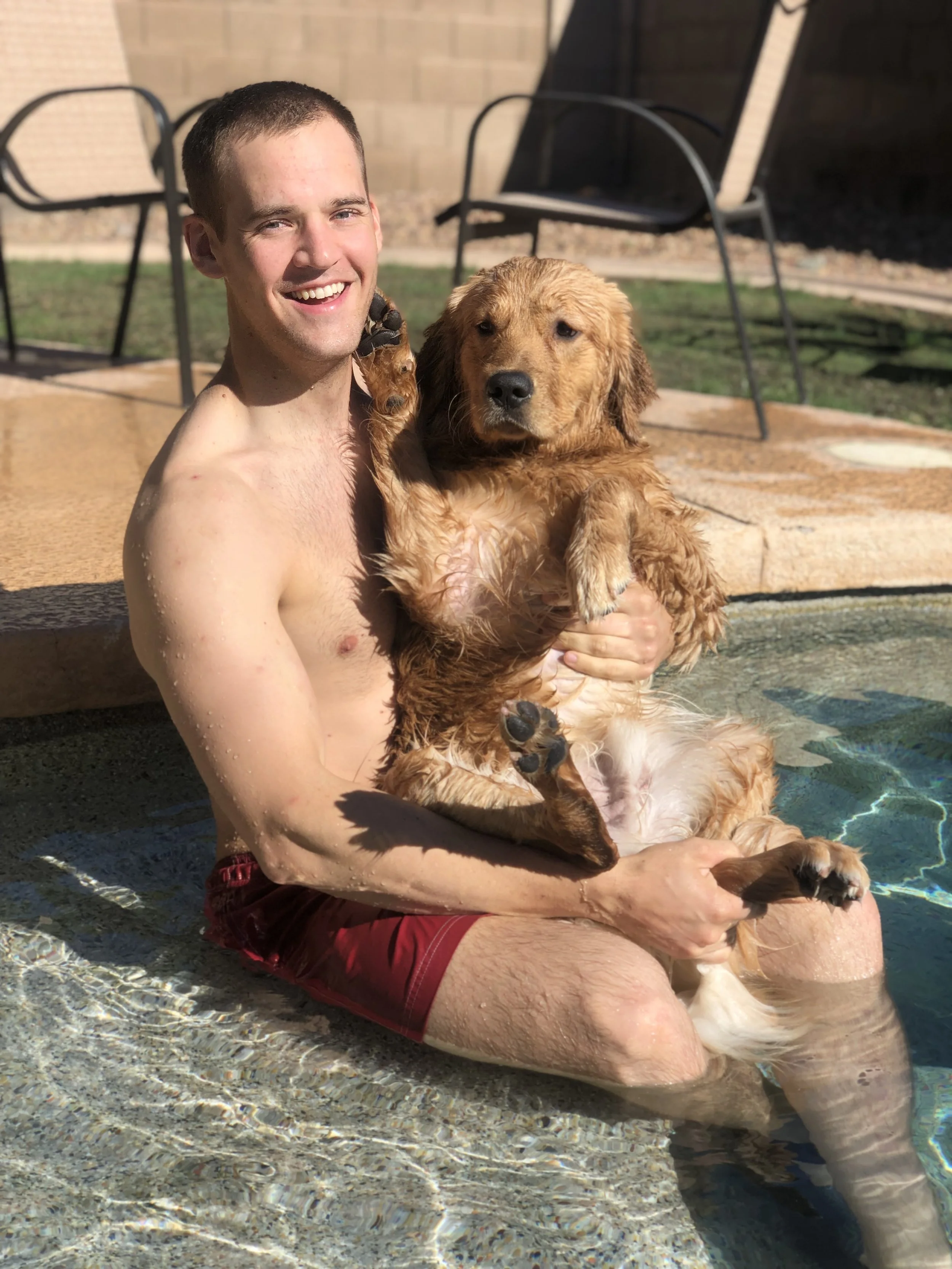A young man with short brown hair smiling while sitting in a pool, holding a wet golden retriever dog, with patio chairs and a brick wall in the background.
