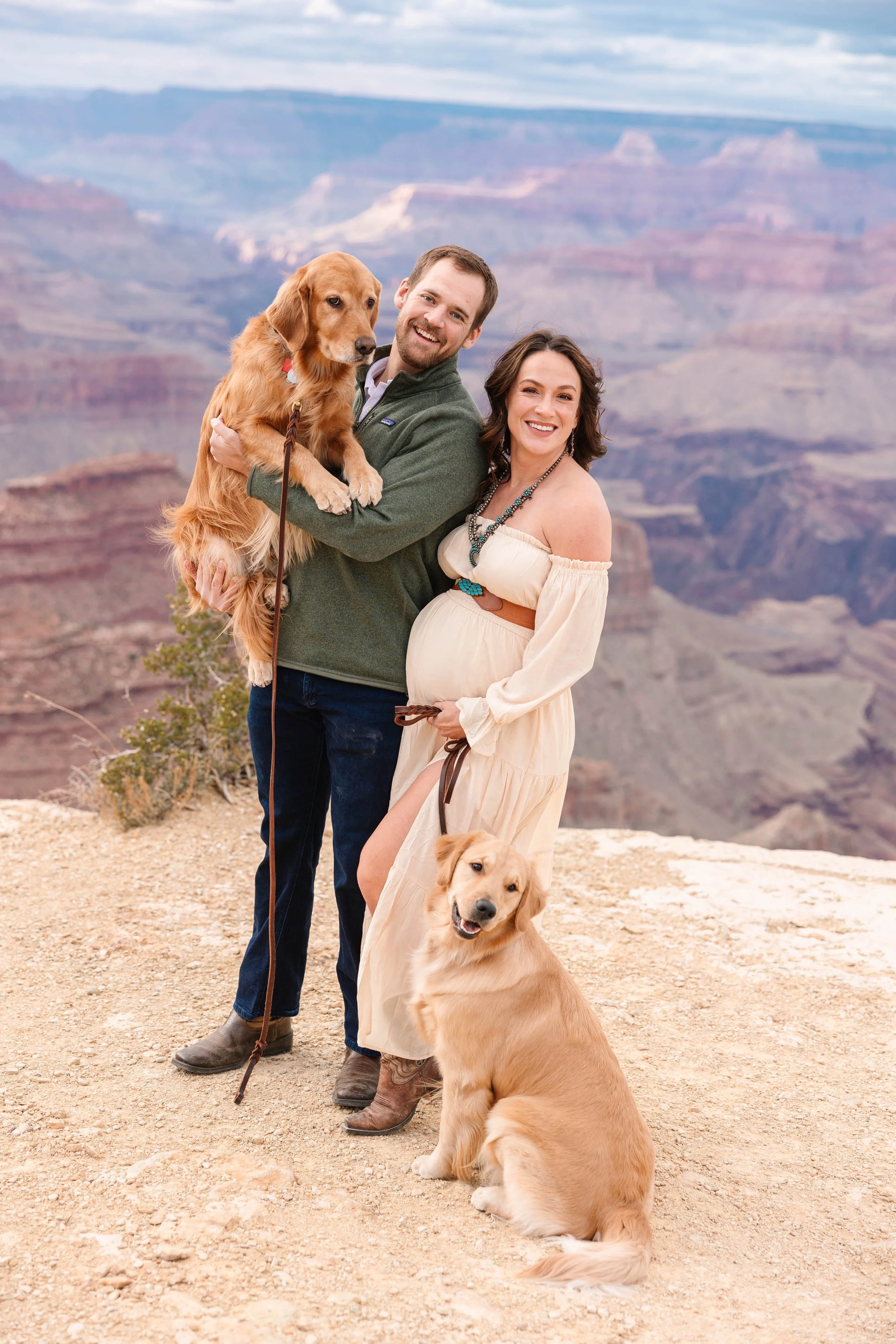 A smiling couple with their golden retriever dogs at the Grand Canyon, with the canyon's layered rock formations and cloudy sky in the background.