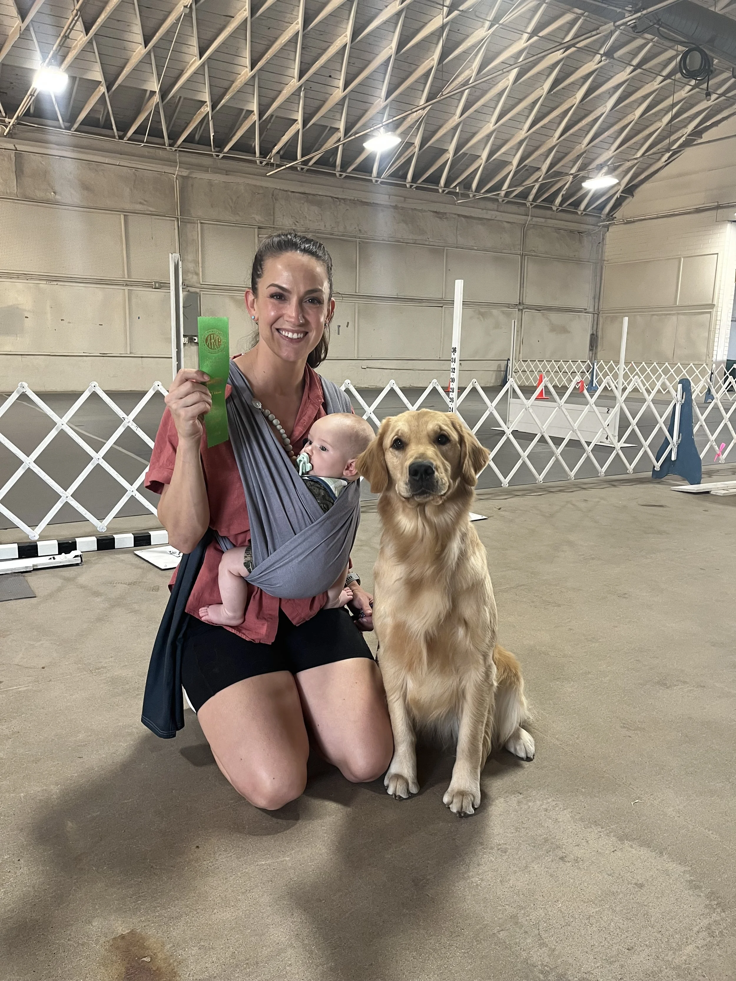 A woman kneeling with a dog and a baby in a baby carrier, inside an indoor dog agility or training area, smiling and holding a green ribbon, with agility equipment and white fencing in the background.