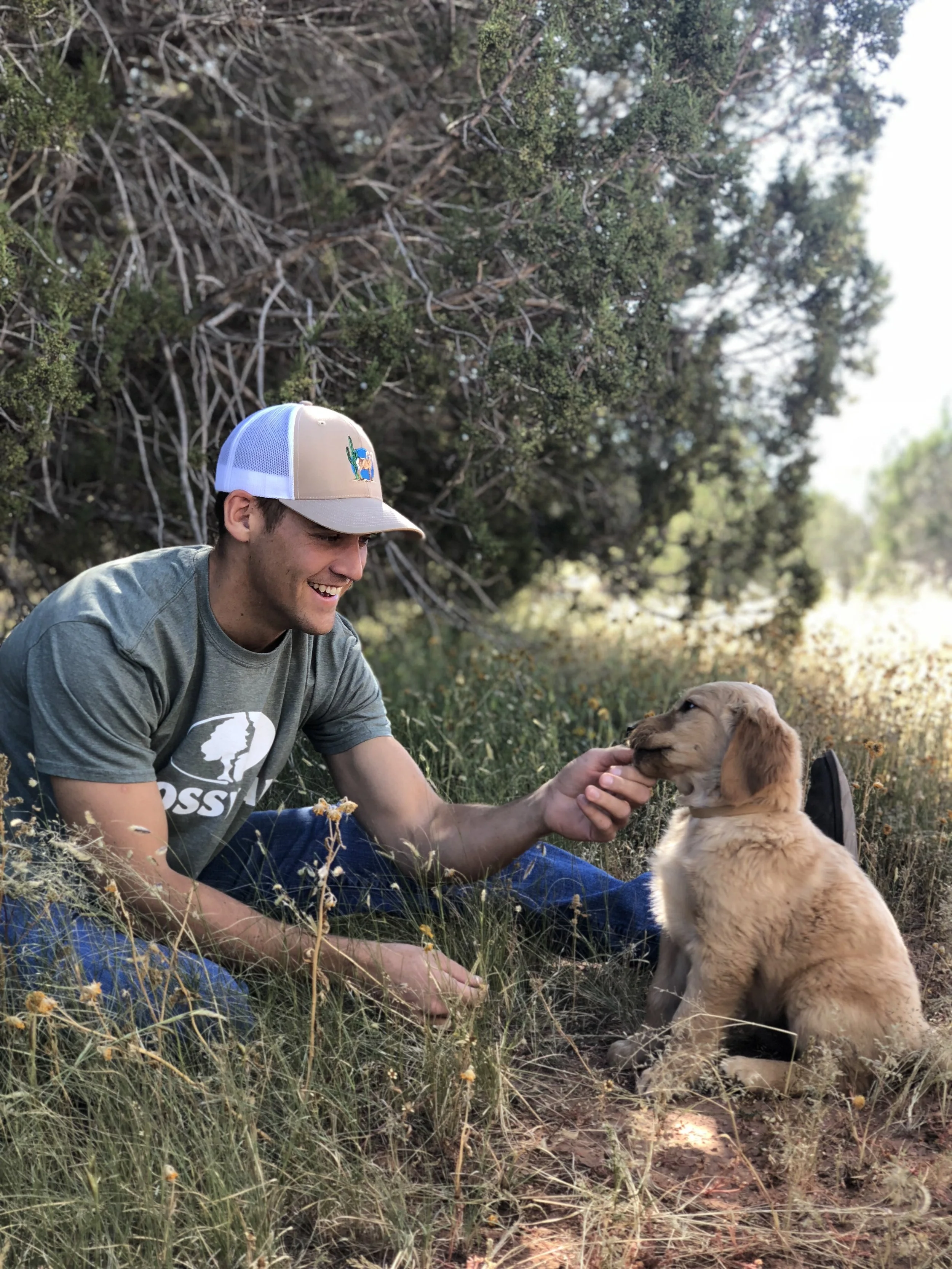 A man wearing a beige and white trucker hat with a cactus patch, sitting outdoors on grass and surrounded by shrubbery, smiling as he interacts with a golden retriever puppy.