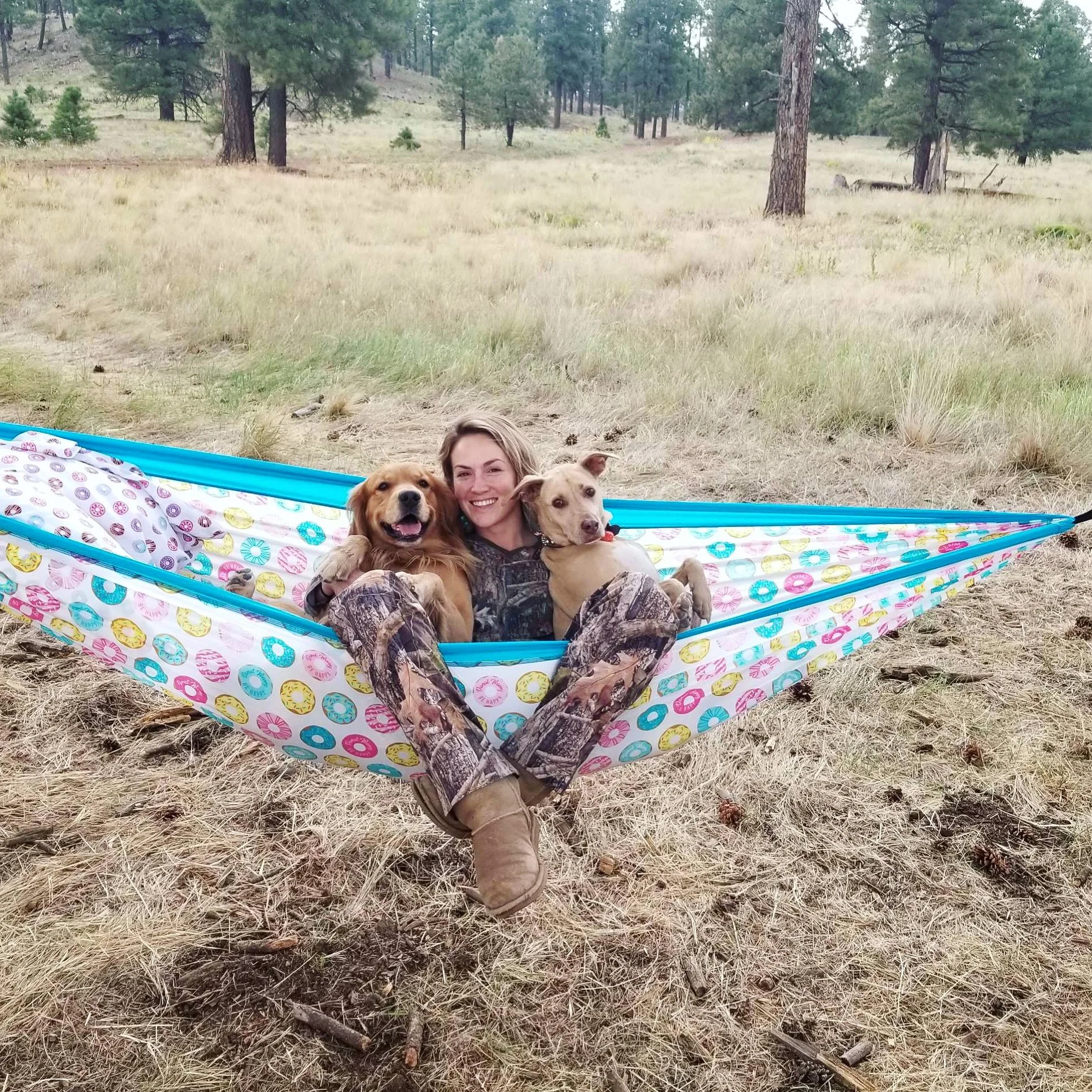 Woman sitting in a colorful hammock with two dogs in a grassy, wooded outdoor area.