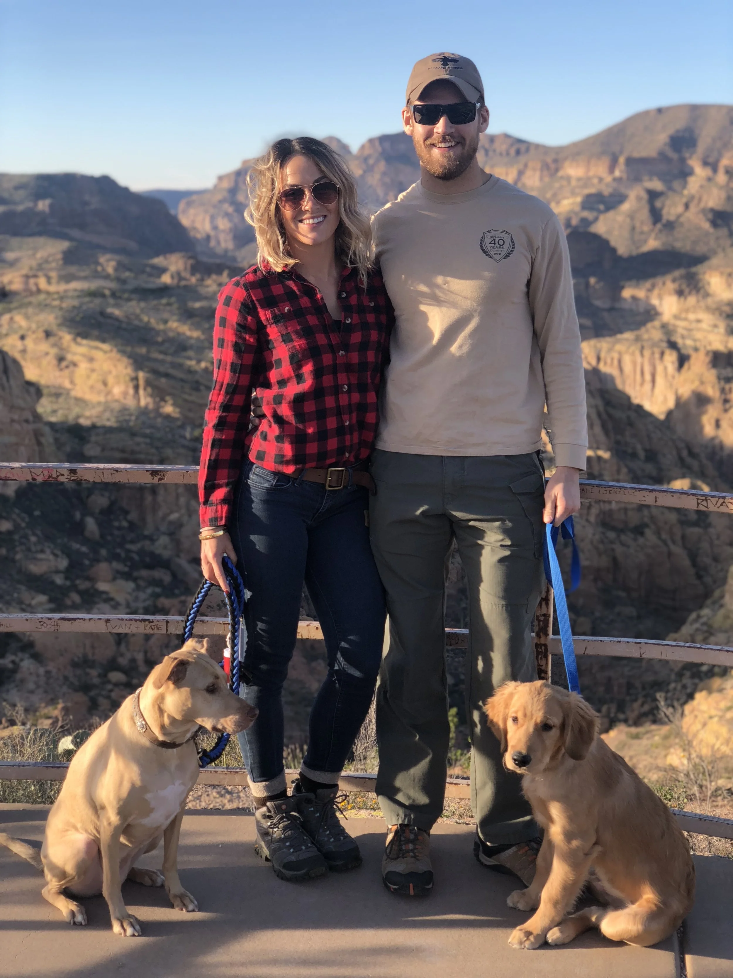 A smiling couple with two Golden Retrievers in a desert landscape with mountains in the background. The woman is wearing sunglasses, a red and black plaid shirt, and jeans, while the man is wearing a beige long-sleeved shirt, a cap, and sunglasses. T