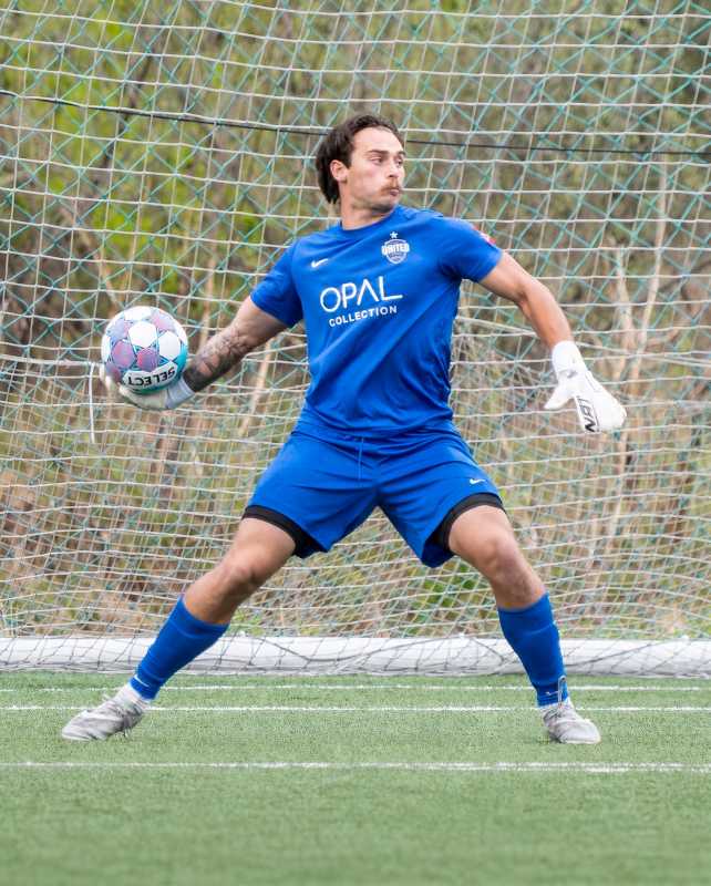 A male soccer goalkeeper in blue uniform making a save with a soccer ball in a goal area.