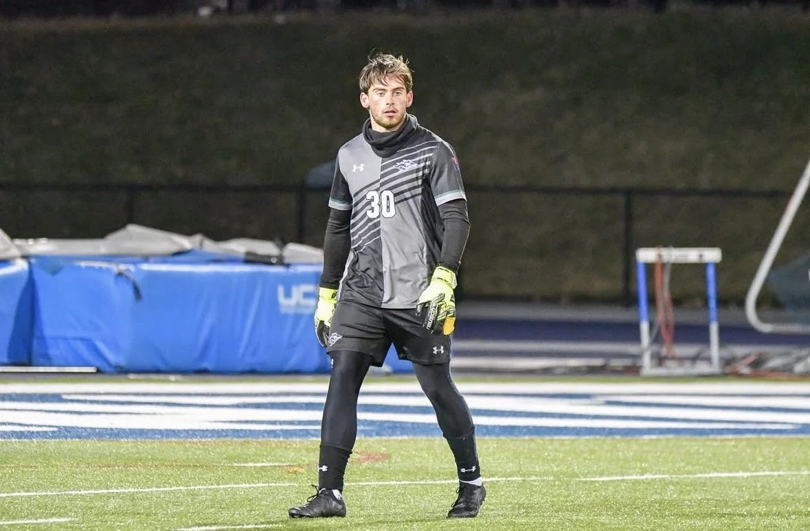A young man dressed as a soccer goalkeeper standing on a field during a game or practice, wearing a black and gray uniform with the number 30, gloves, and black leggings.