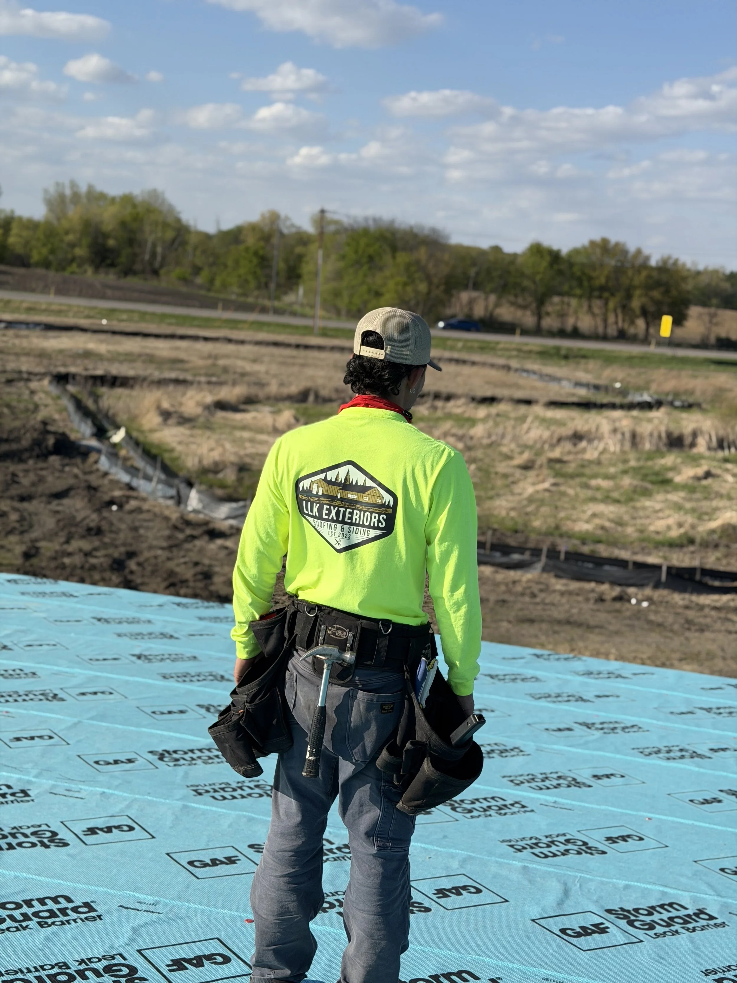 A construction worker in a bright yellow safety shirt and tool belt is standing on a blue ground barrier at a construction site, with a landscape of trees, a road, and a blue sky with clouds in the background.