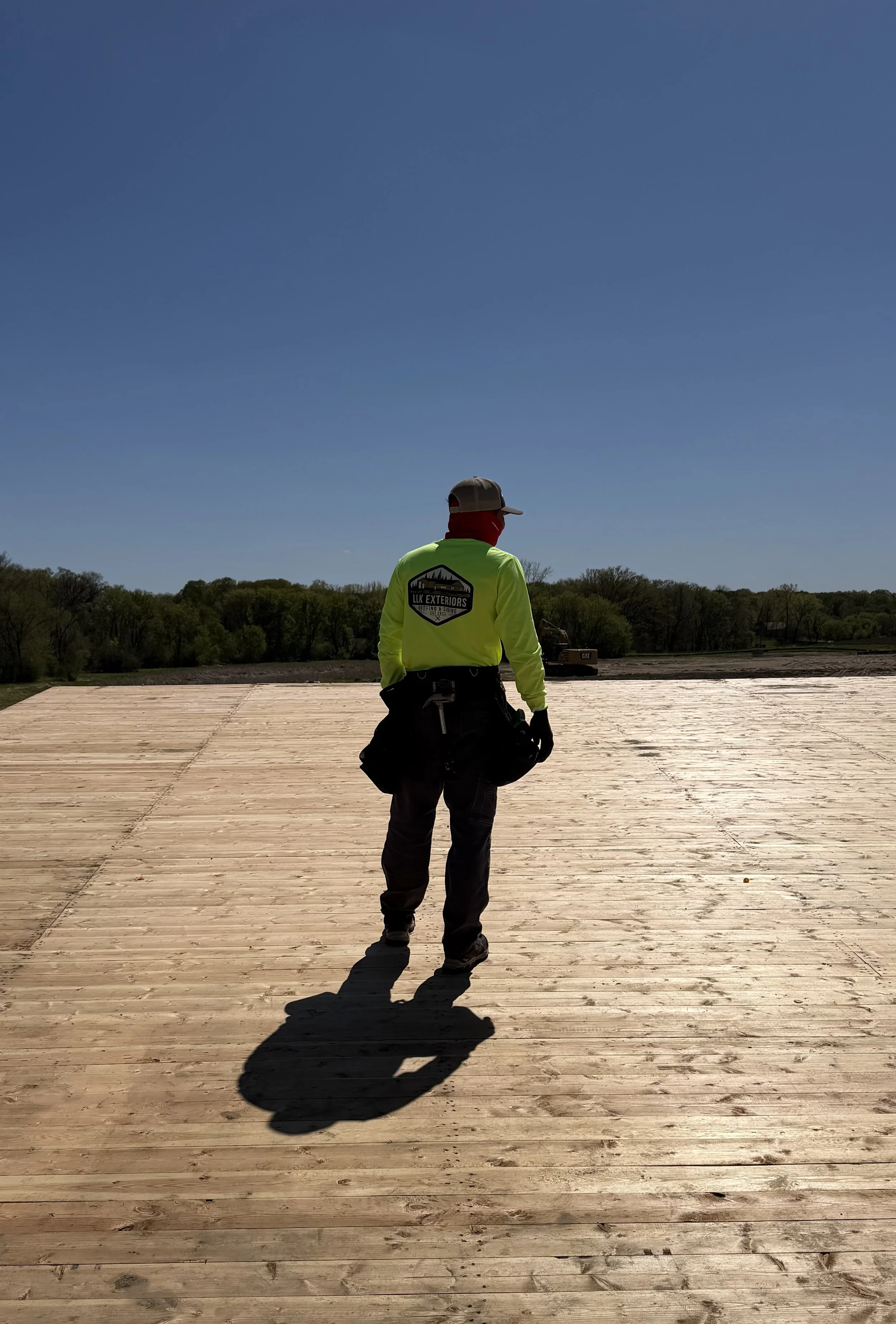 A construction worker standing on a wooden platform with his back to the camera, wearing a bright yellow jacket with a logo on the back, a gray cap, and holding a black bag, under a clear blue sky with a distant tree line.