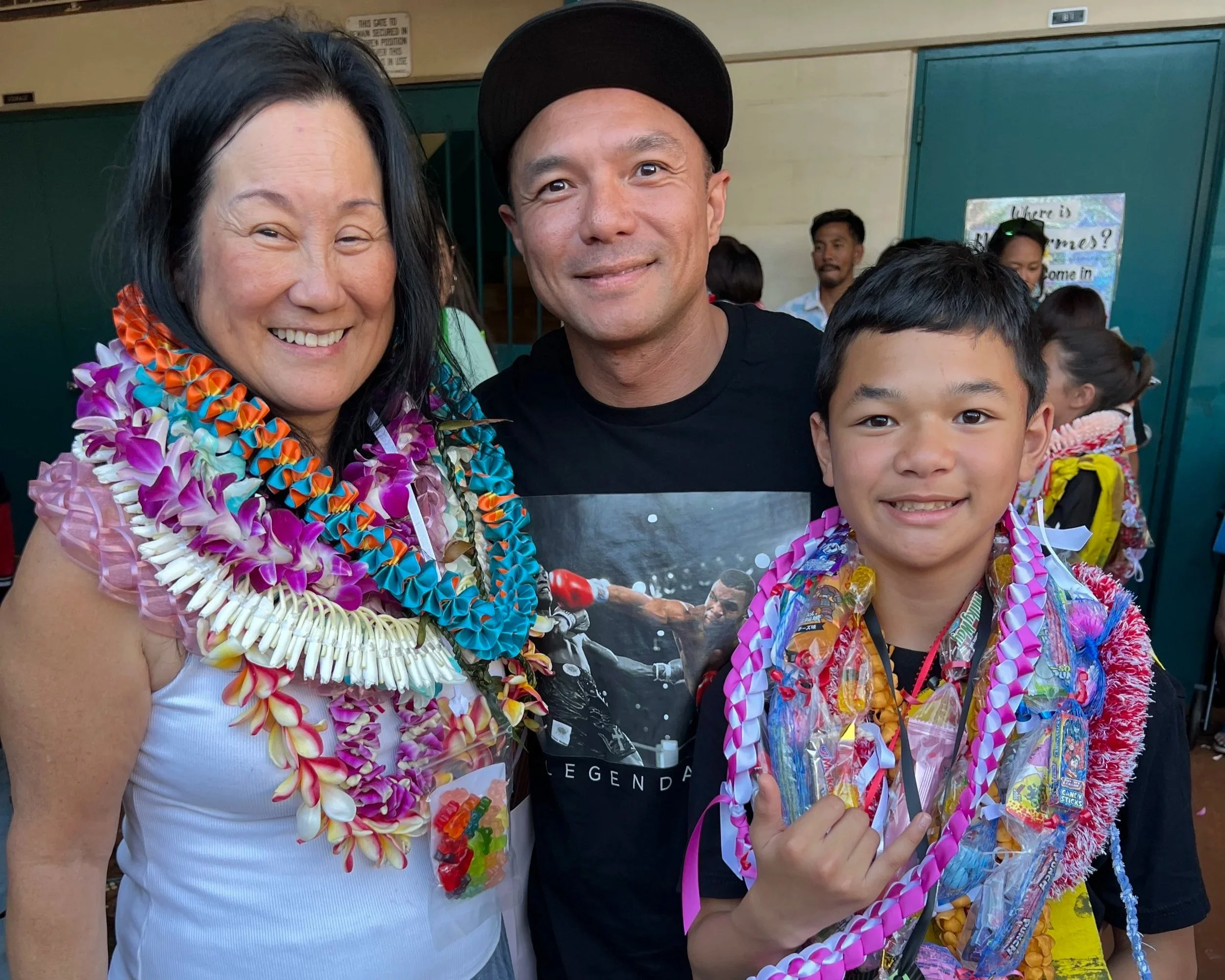 Three people smiling, wearing colorful leis, at a celebration or gathering, with a background of others and a sign on the wall.