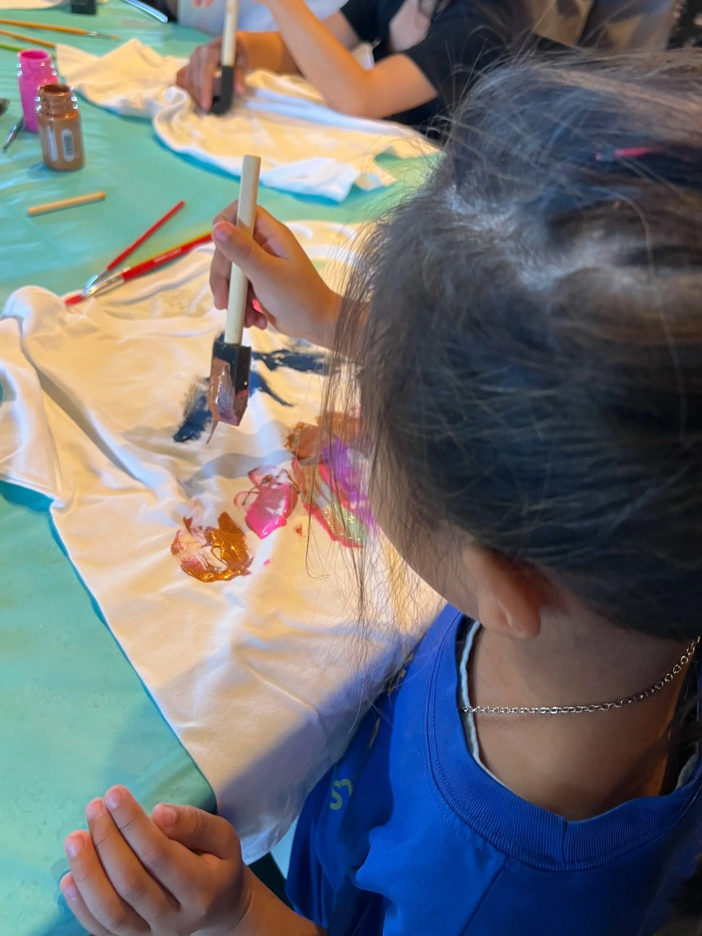 Child painting on a piece of fabric with a paintbrush, surrounded by paint jars and other art supplies on a table.