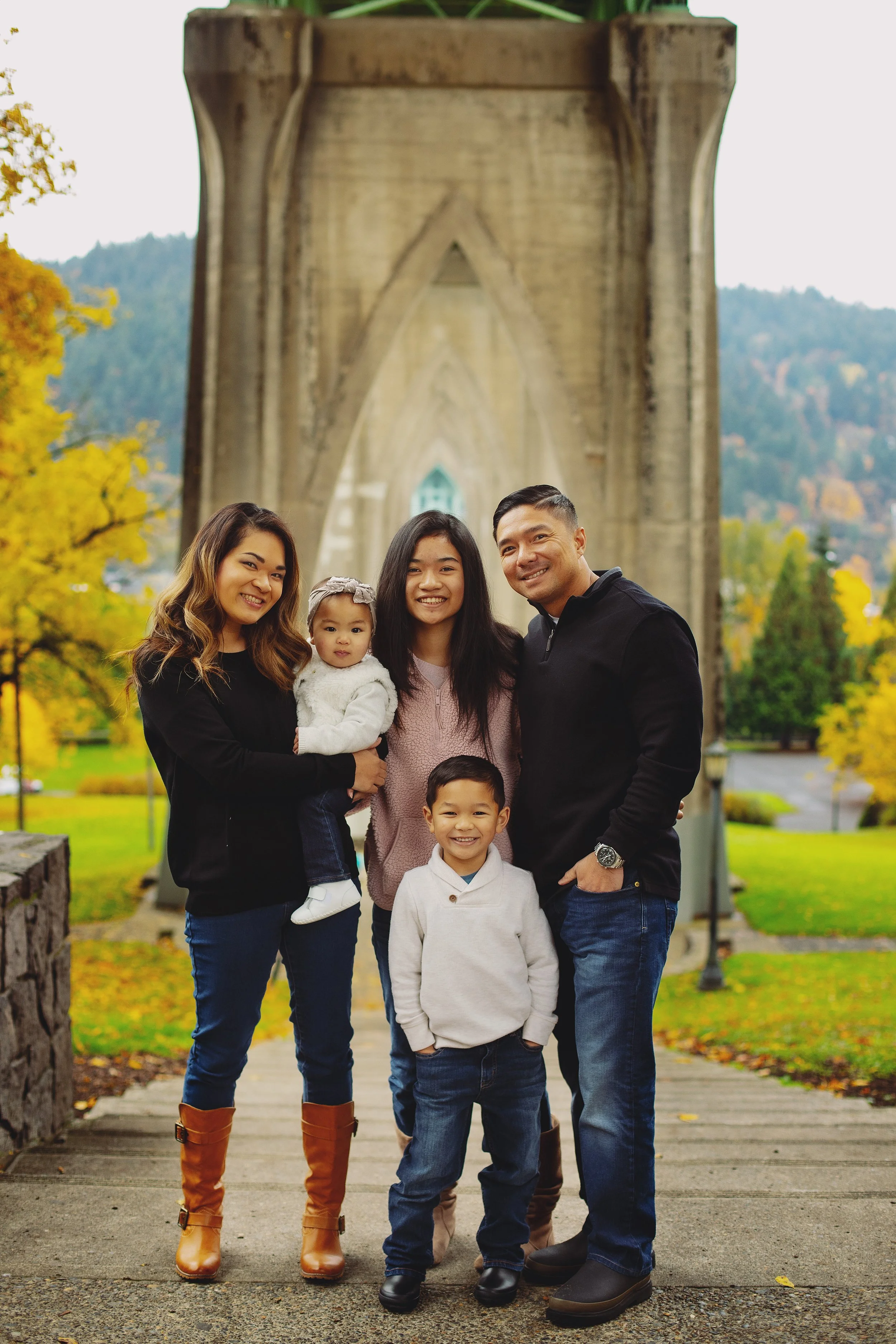 A family of six standing on a wooden pathway in front of a large stone bridge, autumn trees with yellow leaves in the background. The family includes two adults, two children, and two infants. They are smiling and dressed warmly for fall.