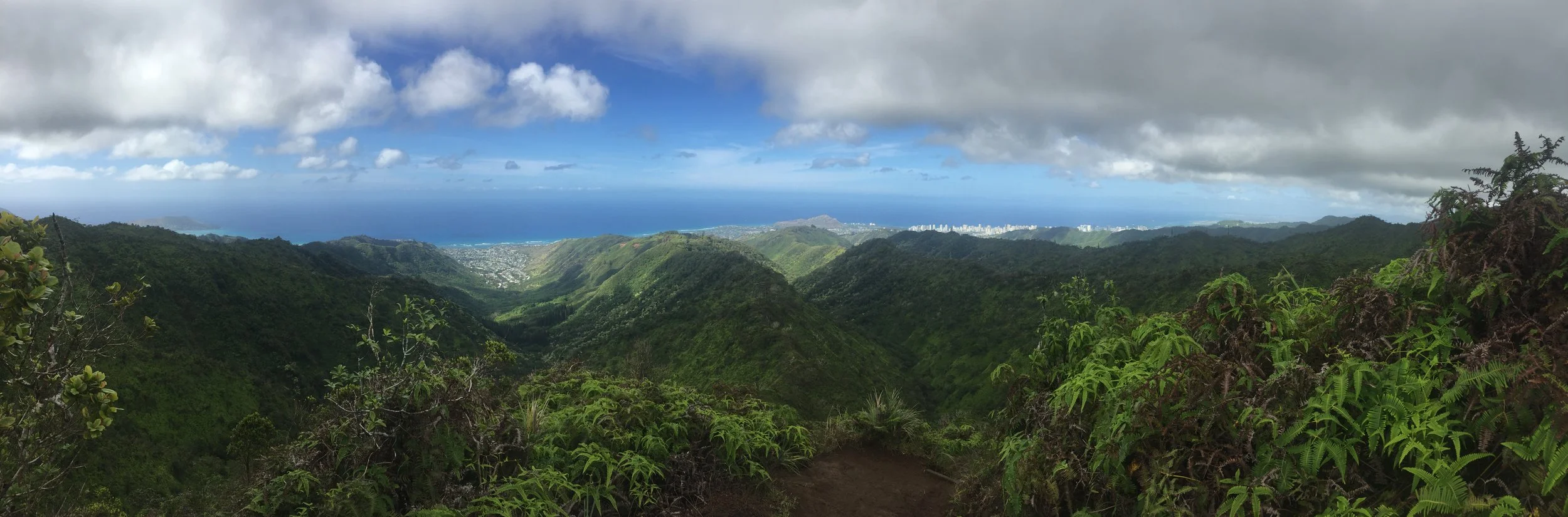 Panoramic view of lush green mountains, with blue ocean and sky in the background, and scattered clouds overhead.