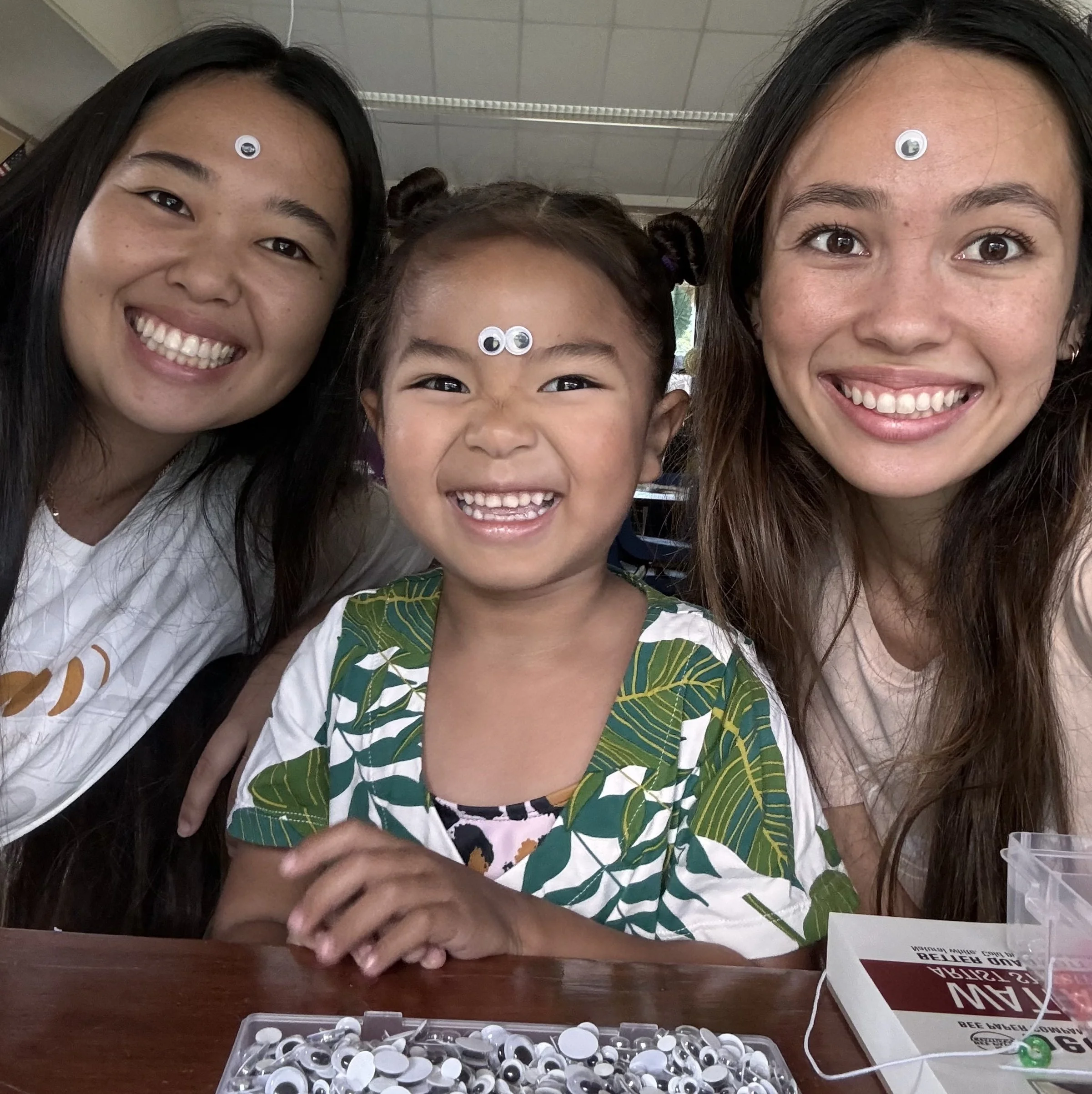 Three smiling women, two with dark hair and one with light brown hair, shirts with bright patterns, wearing googly eyes stickers on their foreheads, sitting at a table with a box of white and black googly eyes, a plastic container, and a book titled 'How to Beat Fear and Anxiety'.