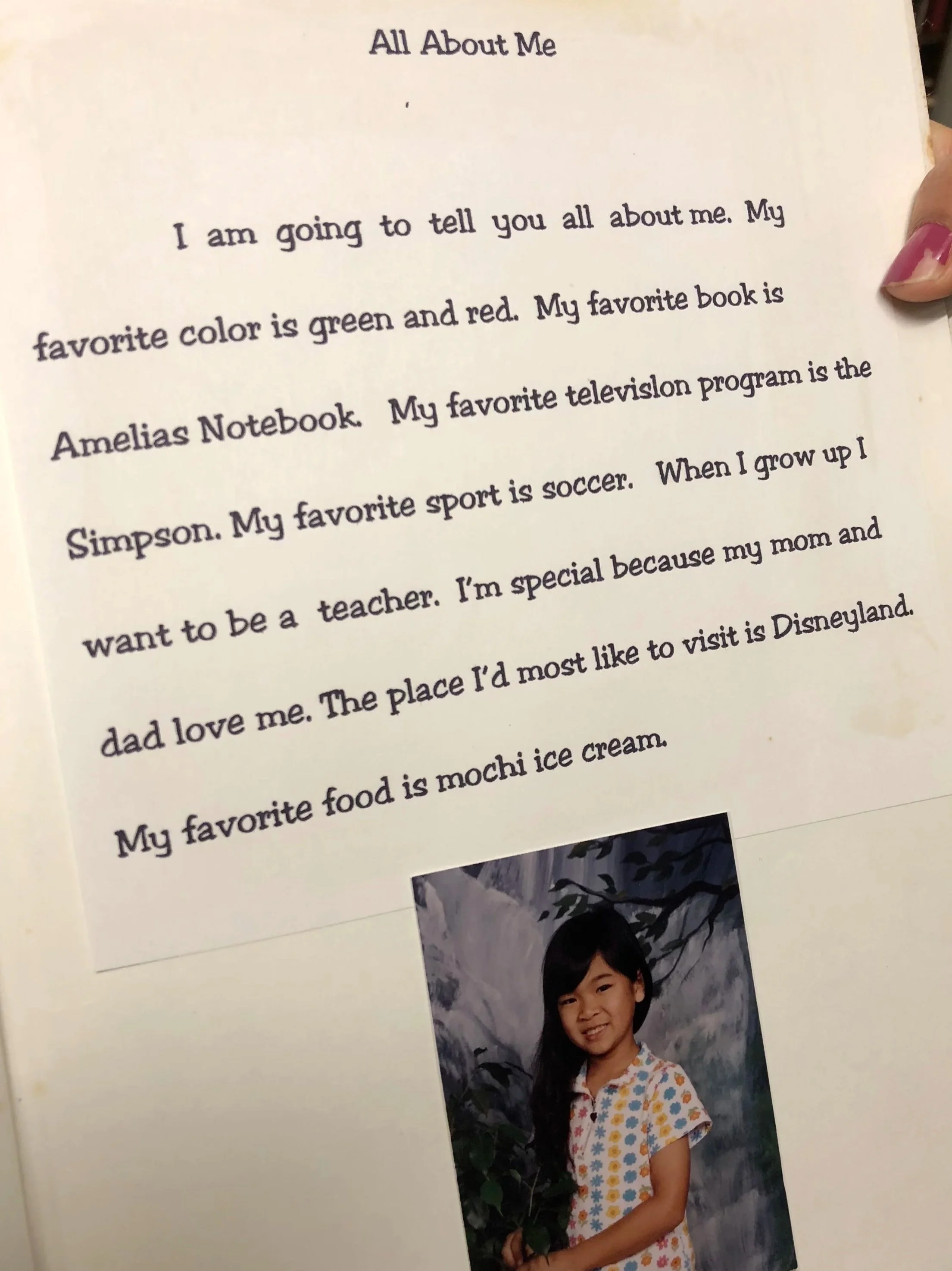 A page from a book titled "All About Me" with text about a girl, and a photograph of a young girl with long dark hair wearing a colorful flowered shirt, standing outdoors against a background of mountains and trees.