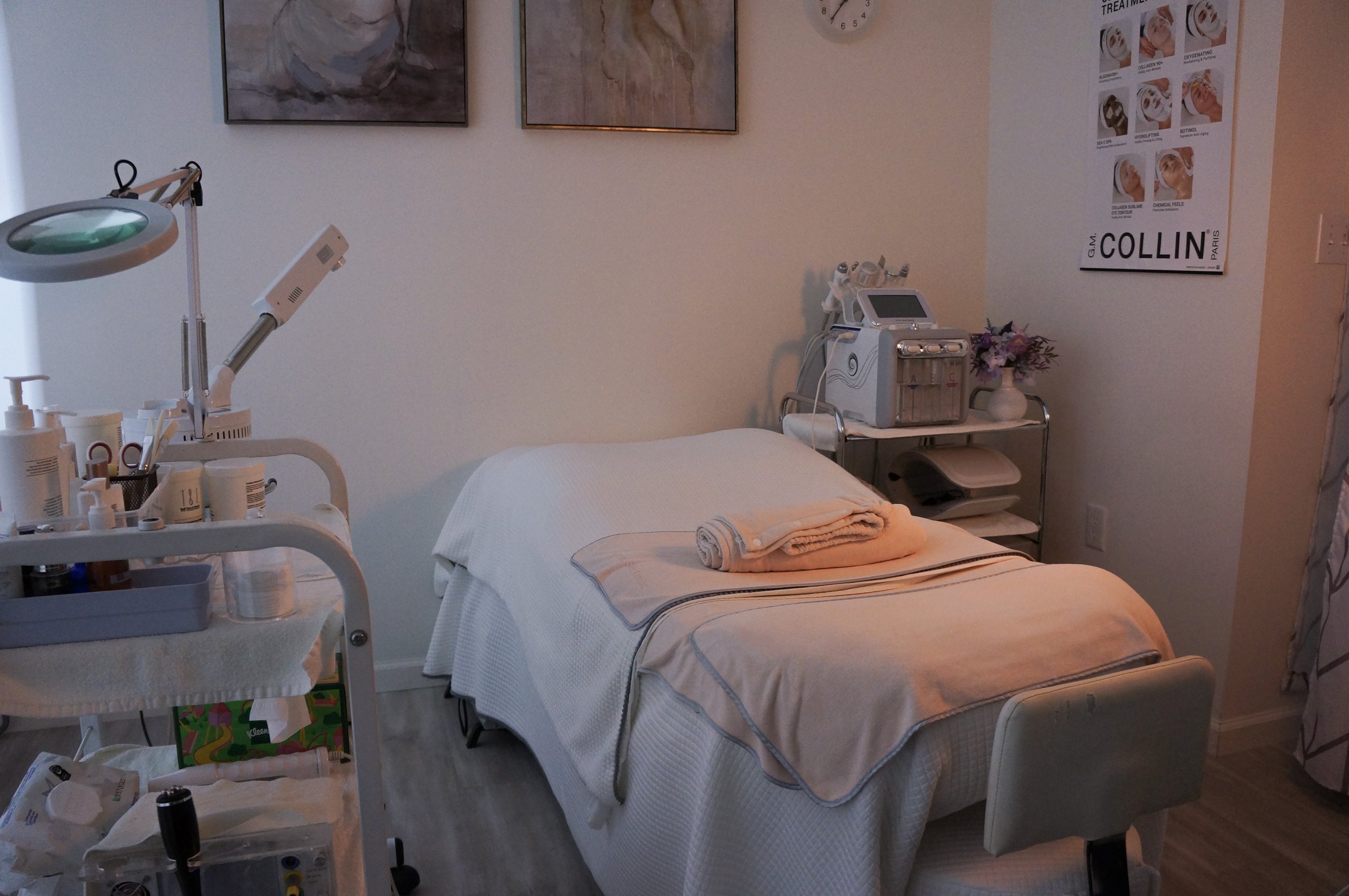 A medical treatment room with an examination bed covered with white linens and a folded towel. To the left, there's a medical cart with various supplies and medical equipment. On the right, there's another cart with a vase of flowers, a medical device, and several other machines. The wall in the background has framed artwork and a poster detailing facial treatment procedures. The room has soft lighting and a clean, organized appearance.