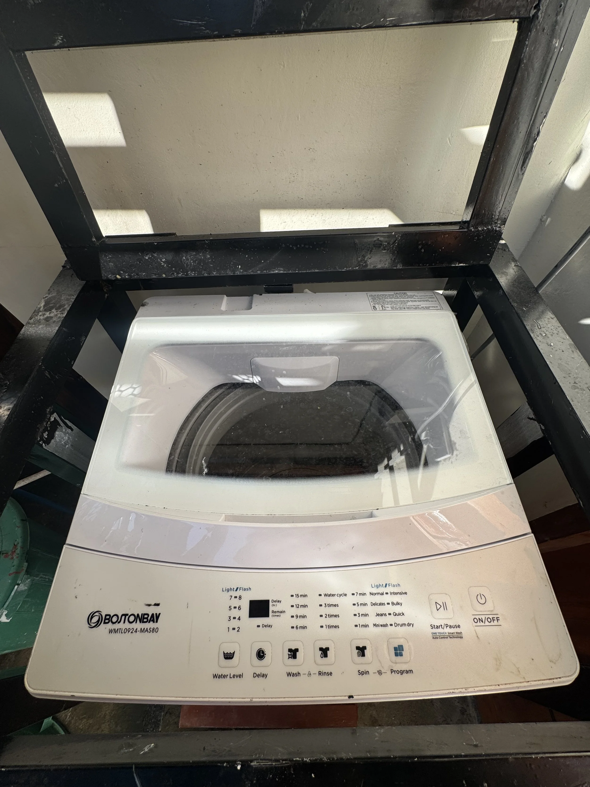 A top-loading washing machine with control buttons and labels on the front panel, situated on a black metal stand, with a white wall in the background.