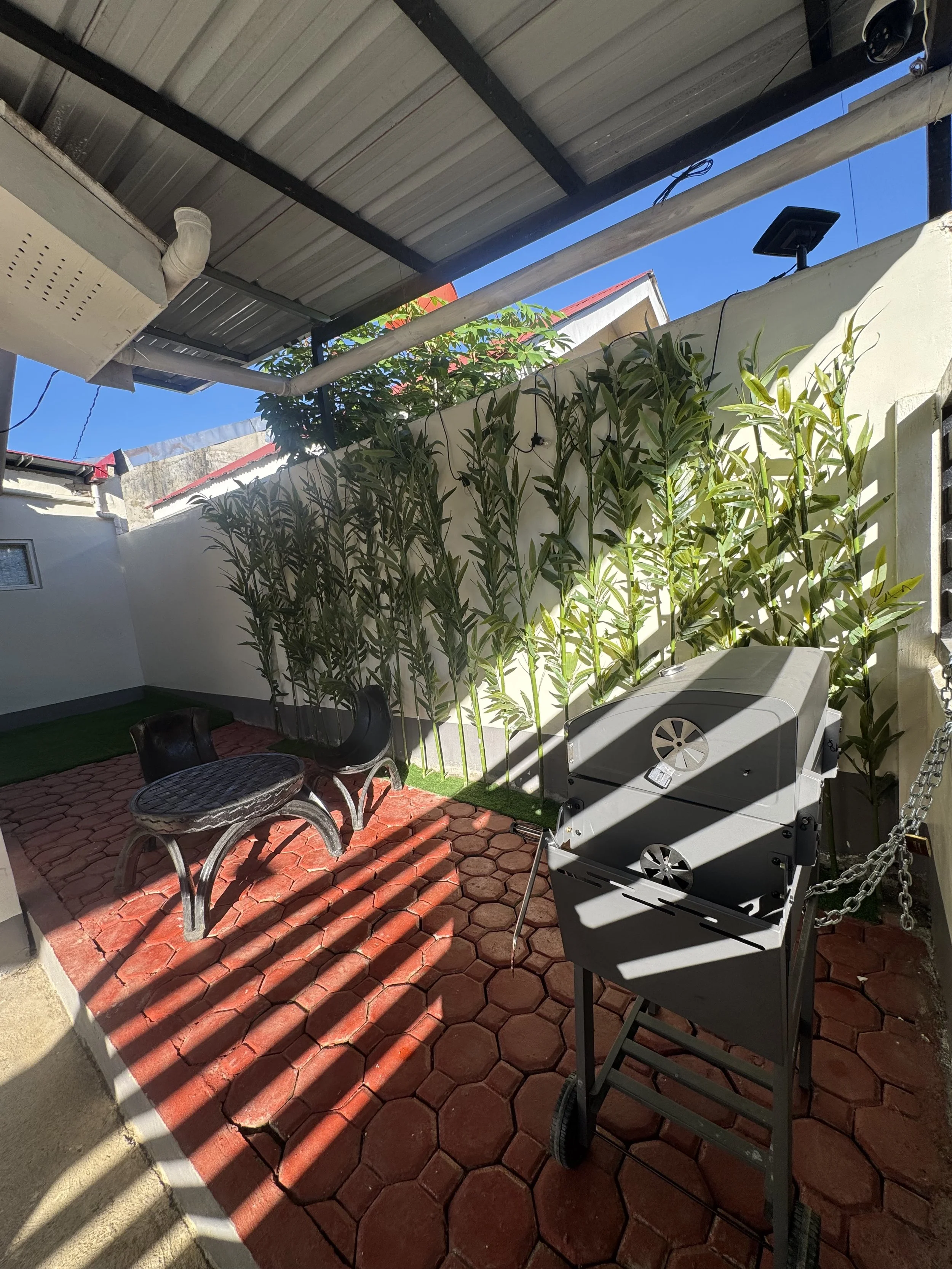 Patio with red brick flooring, outdoor grill, two chairs, a round table, potted plants, and a white wall. Part of the roof and some electrical wires are visible, with a bright blue sky in the background.