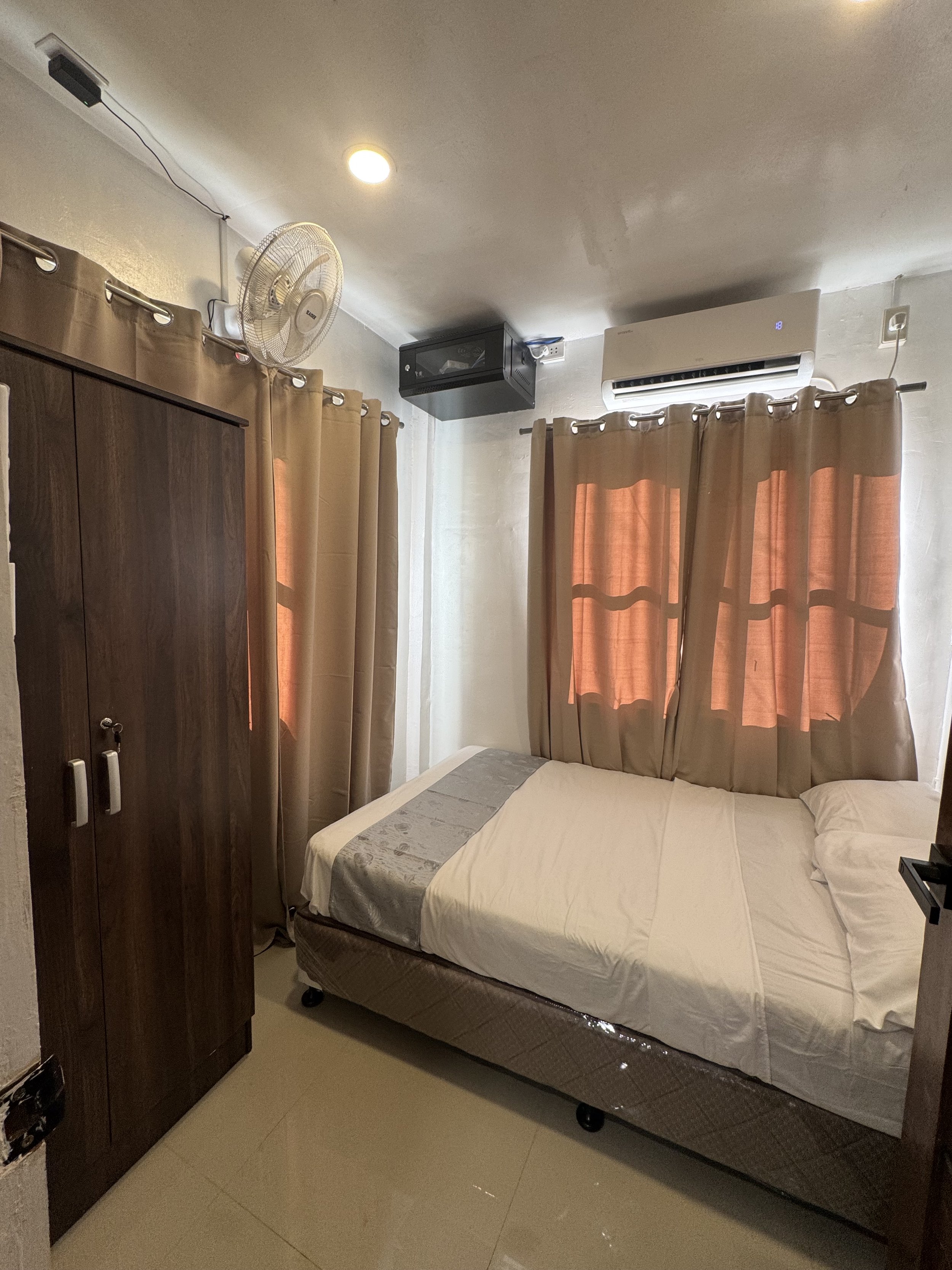 A bedroom with beige curtains, a bed with a white mattress, a headboard, and a wardrobe, with a wall-mounted fan and air conditioner visible in the room.