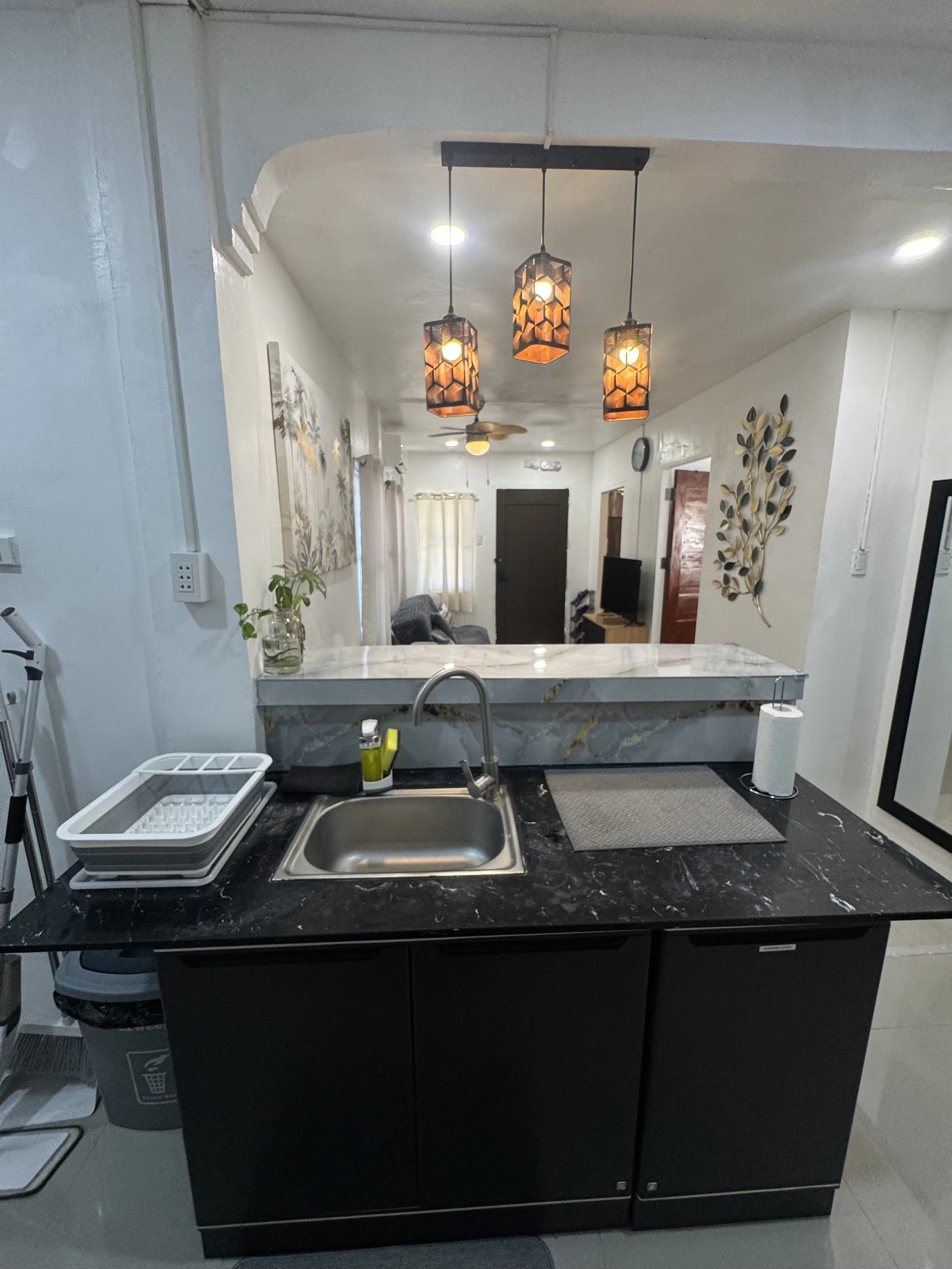 View of a kitchen island with a marble countertop, a sink, and kitchen items, with a living room in the background illuminated by ceiling lights and a three pendant light fixture.