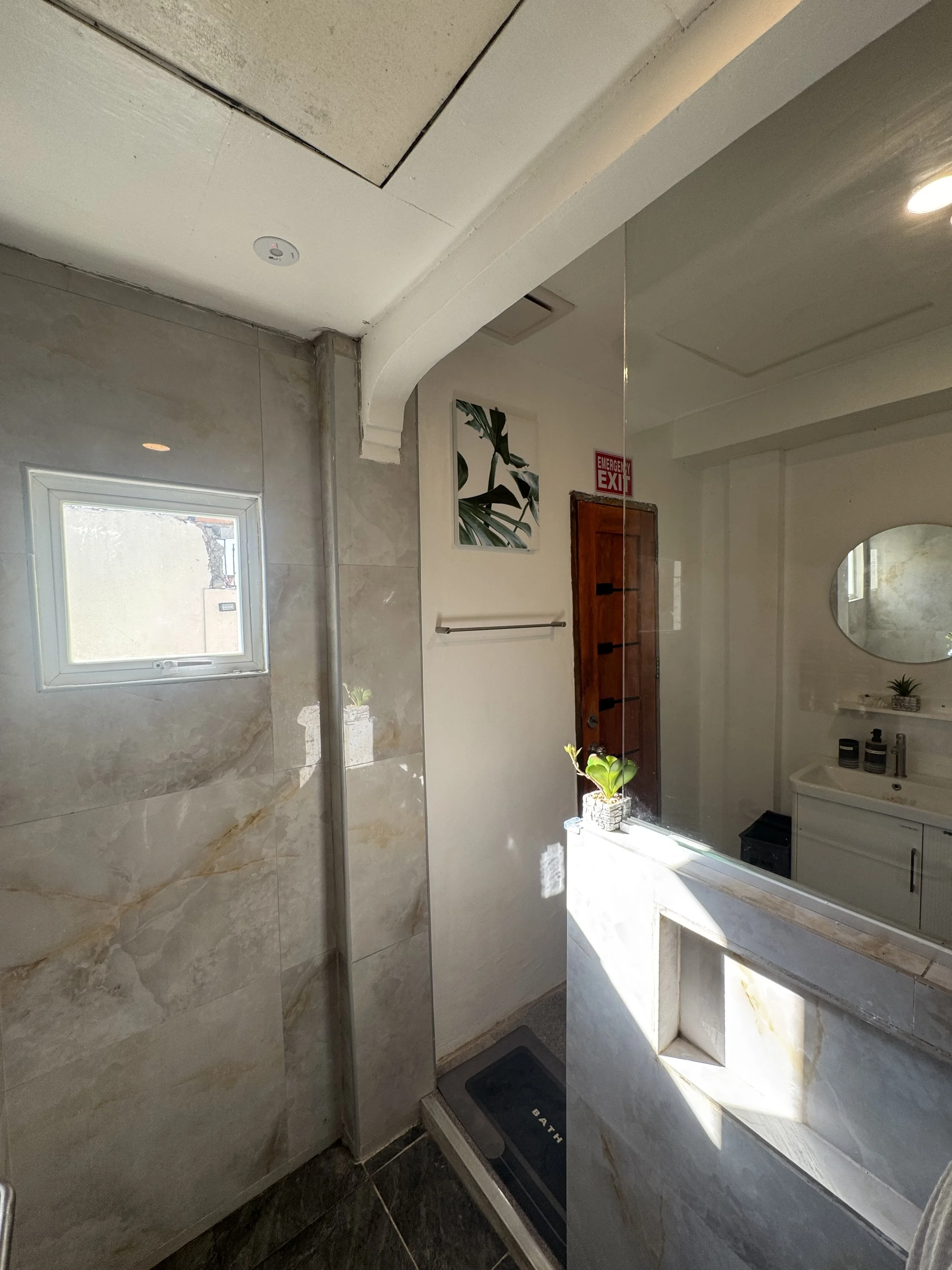 A bathroom with a small window, a plant on a shelf, a mirror, a sink, and a cabinet. The wall tiles are marble-like, and there is an emergency exit sign above a door. Sunlight is entering through the window.