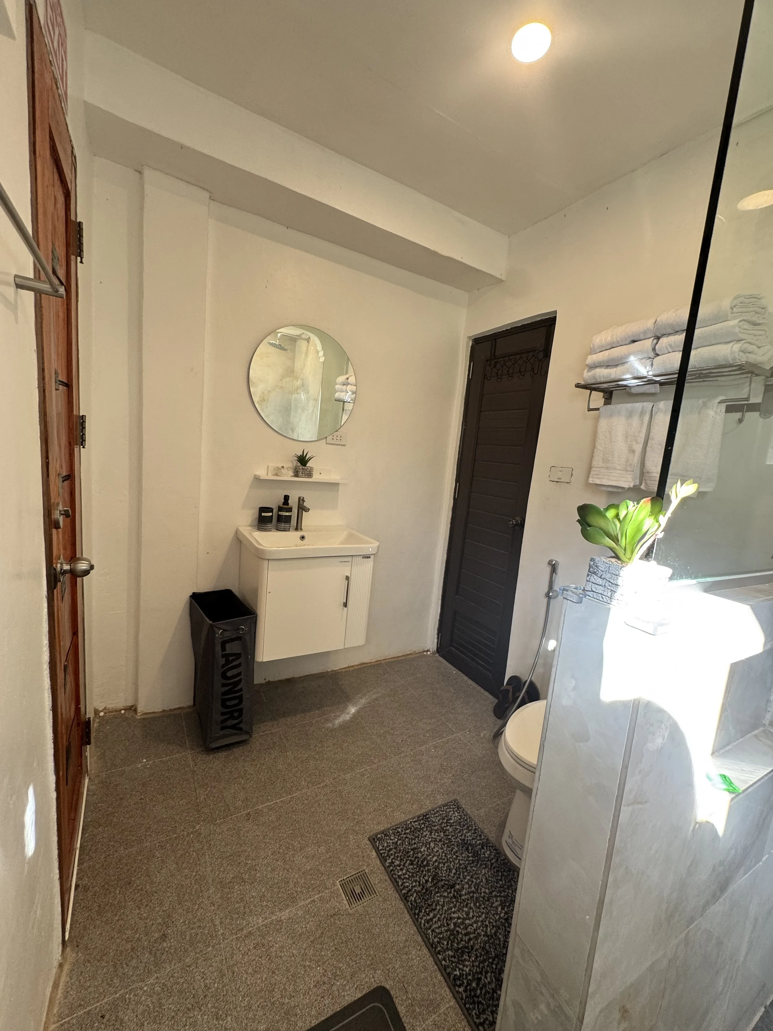 A small bathroom with a white sink, mirror, a black door, and a white water heater. Towels are neatly rolled on a rack, and there is a potted plant near the water heater. The floor is covered with tile, and a black trash bin is placed under the sink.