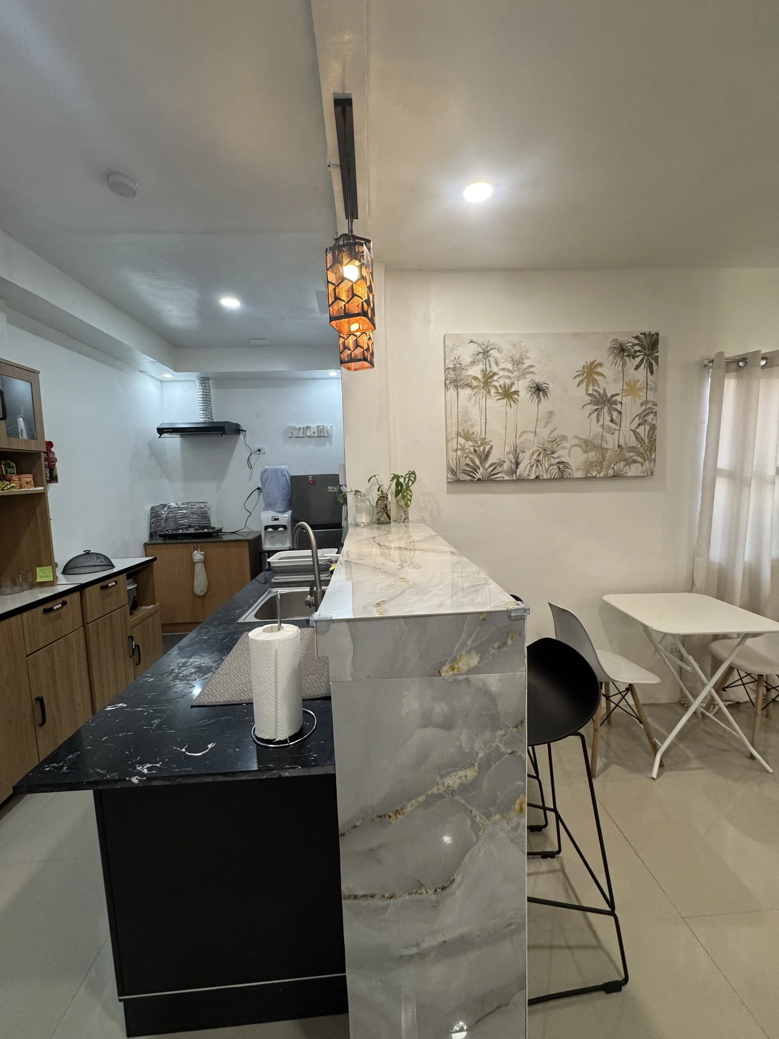 Modern kitchen with a marble island, black and wood cabinets, wall art of palm trees, and seating area with white and black chairs.