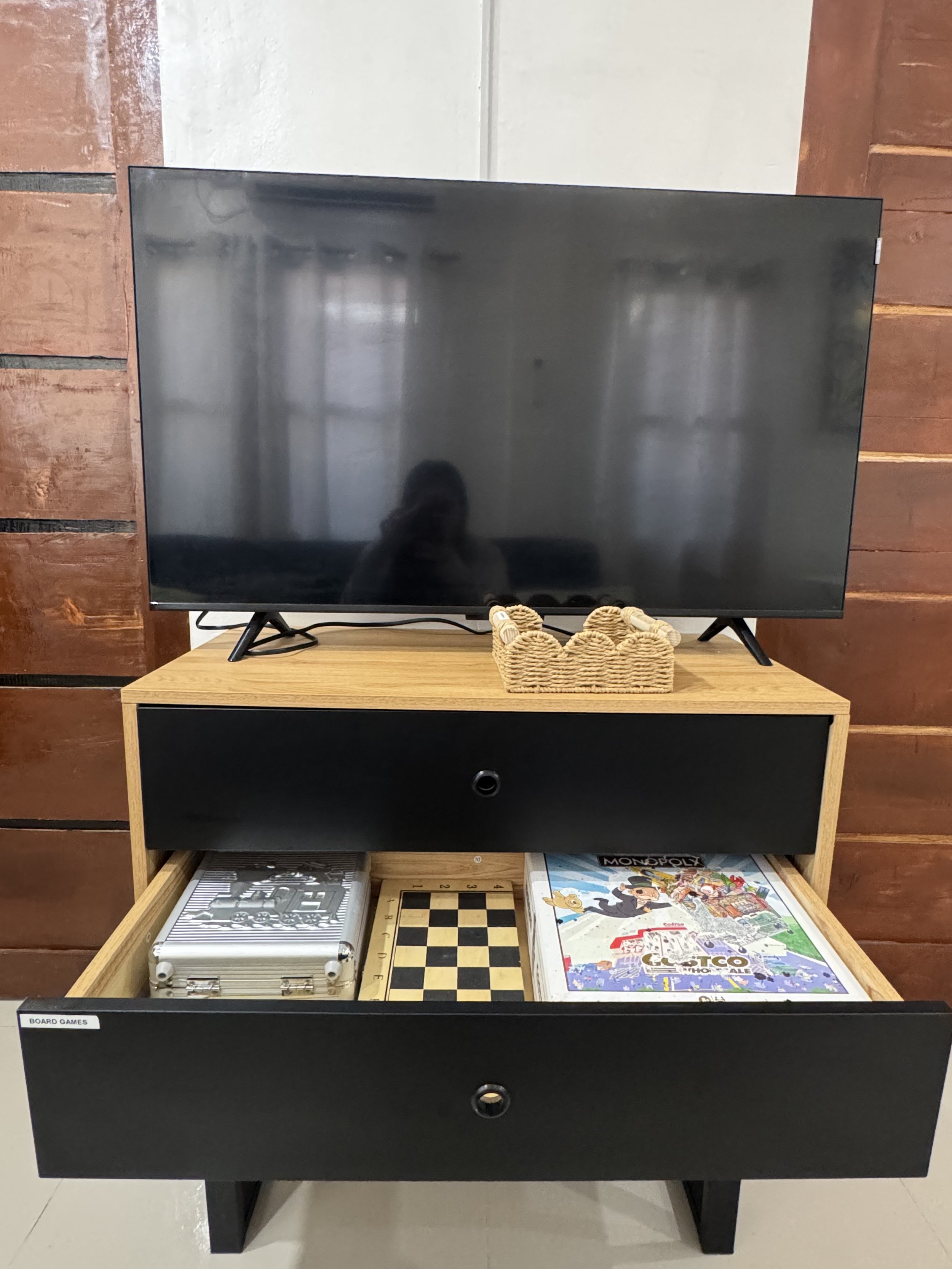 A black and wood-colored table with an open drawer containing board games such as chess, Monopoly, and a connector game. A television is placed on top of the table, and a wicker basket with rope handles is in front of it. The background features dark