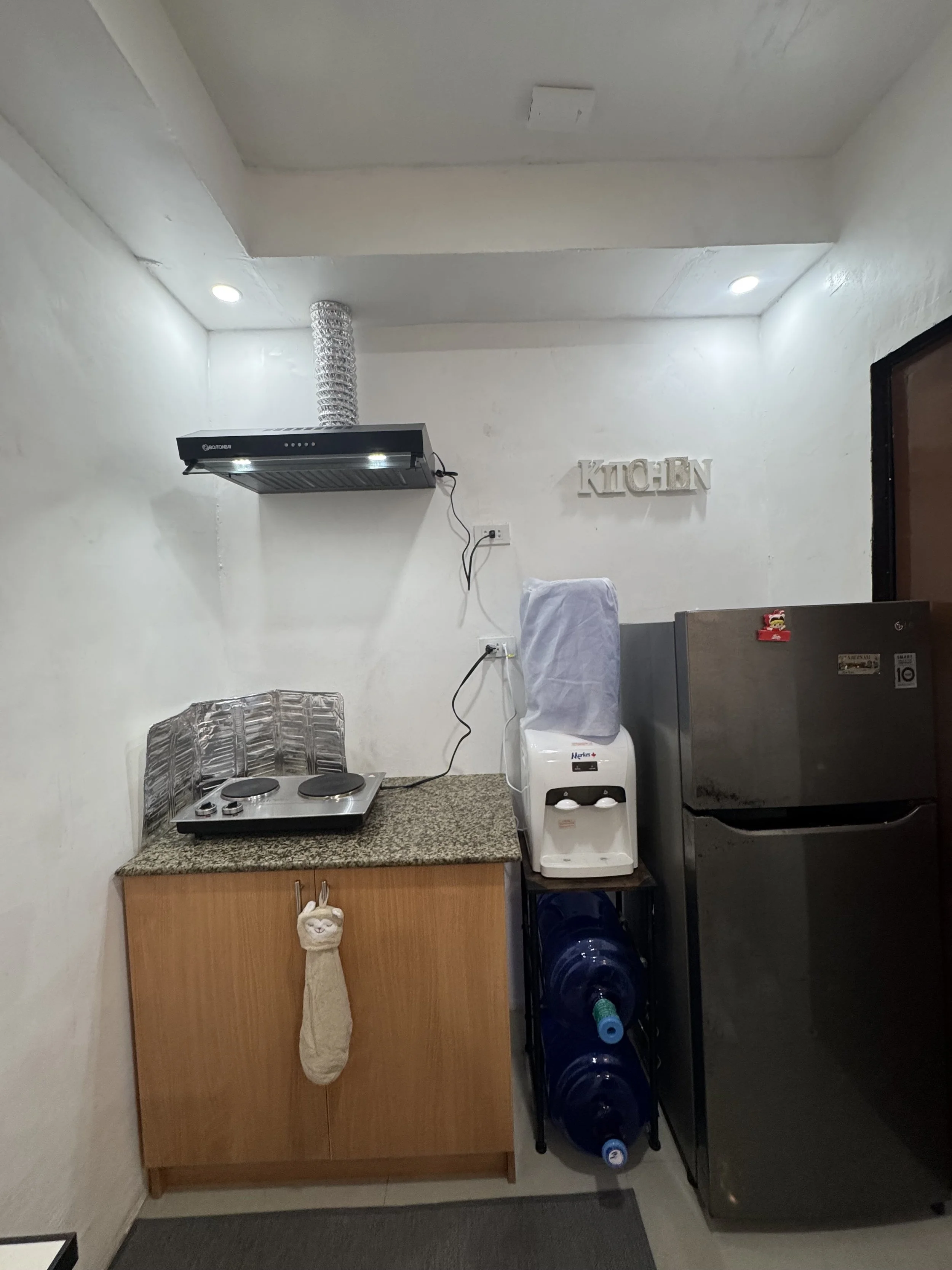 A small kitchen area with a granite countertop, a wooden cabinet with a llama-shaped hanging decoration, a stove with two burners, a water dispenser, a water bottle rack, a refrigerator with magnets, and a water filter under the dispenser. There is a