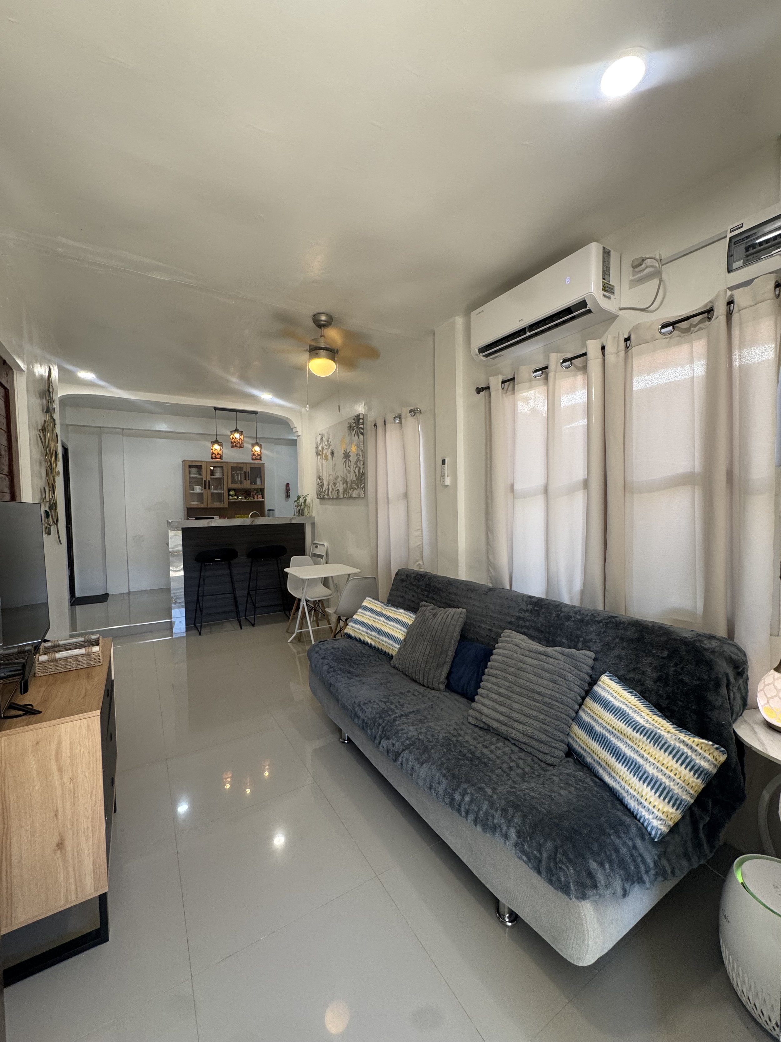Interior view of a living room with a gray sofa, decorative pillows, cream curtains, a wall-mounted air conditioner, a ceiling fan, and a kitchen area in the background with hanging pendant lights and a cabinet.